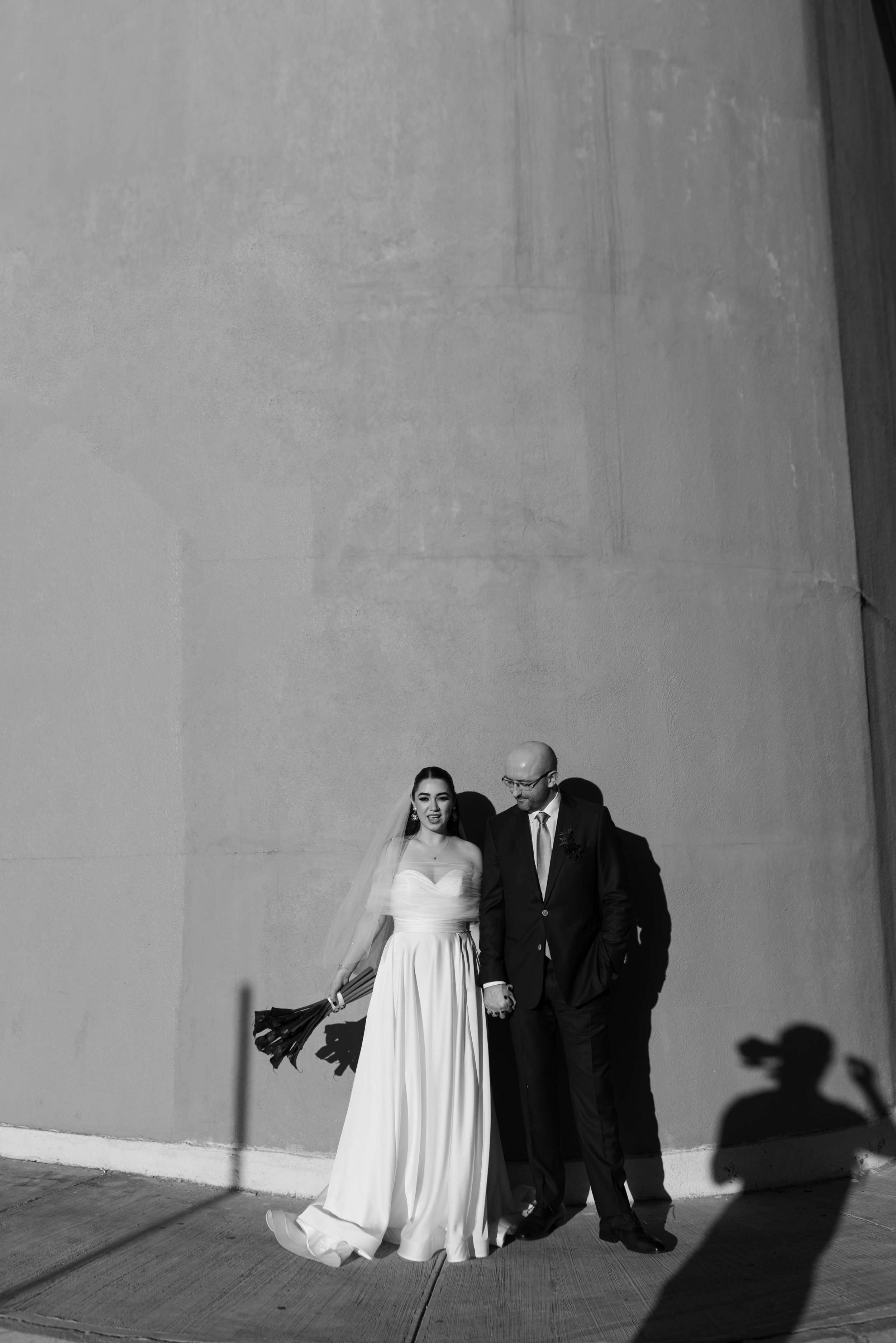 Boda con novia vestida de blanco y novio en traje negro, ambos de pie juntos en la acera frente a un muro.
