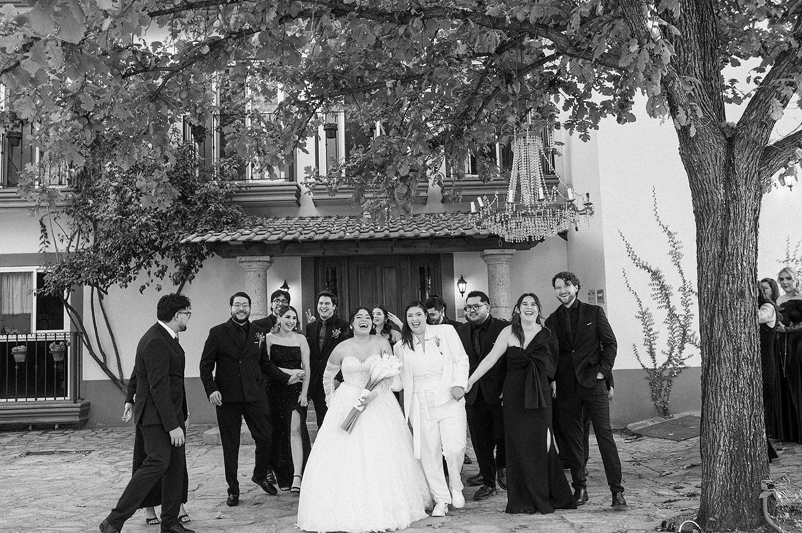 Grupo de personas en celebración de boda, sonriendo y caminando juntos frente a una casa con árbol y candelabros.