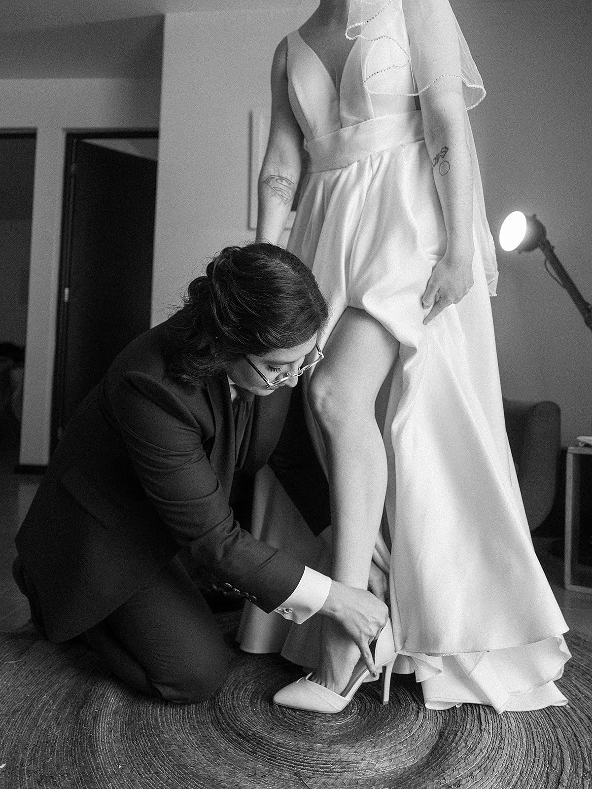 Una mujer con gafas arregla el zapato de un vestido de novia en una habitación. La escena destila atención y detalle.