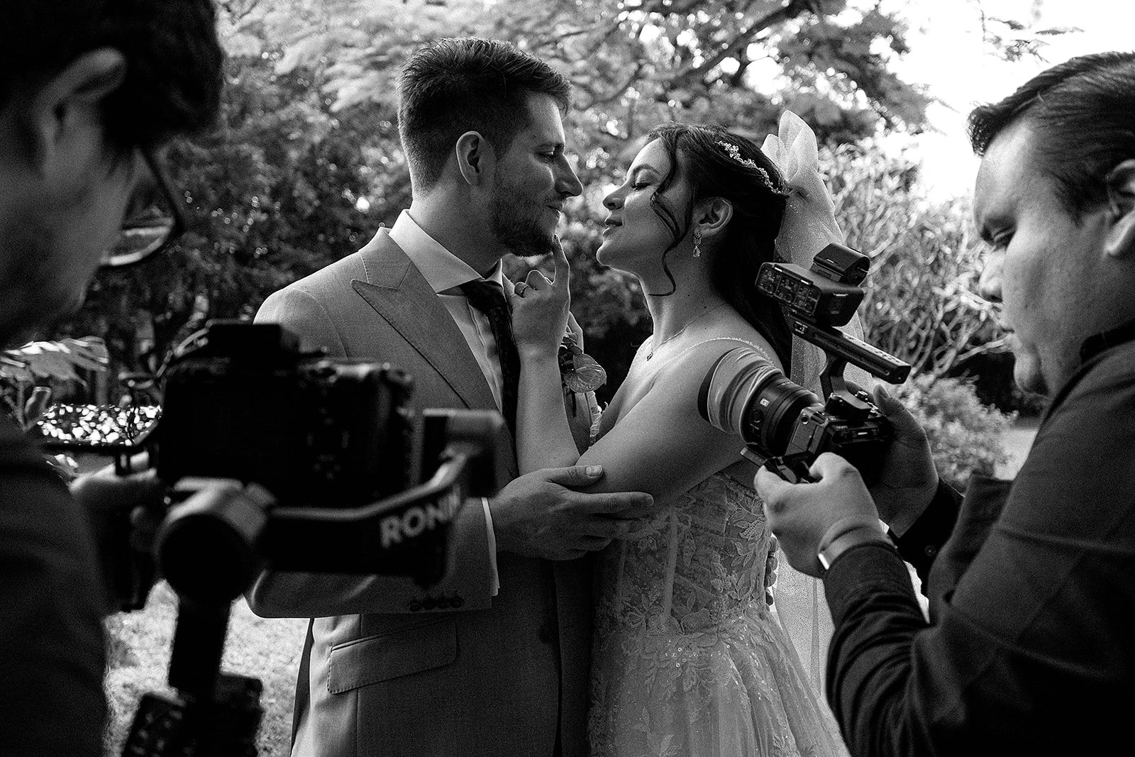 Una pareja se está casando en una ceremonia al aire libre, rodeados de árboles, con fotógrafos capturando el momento.