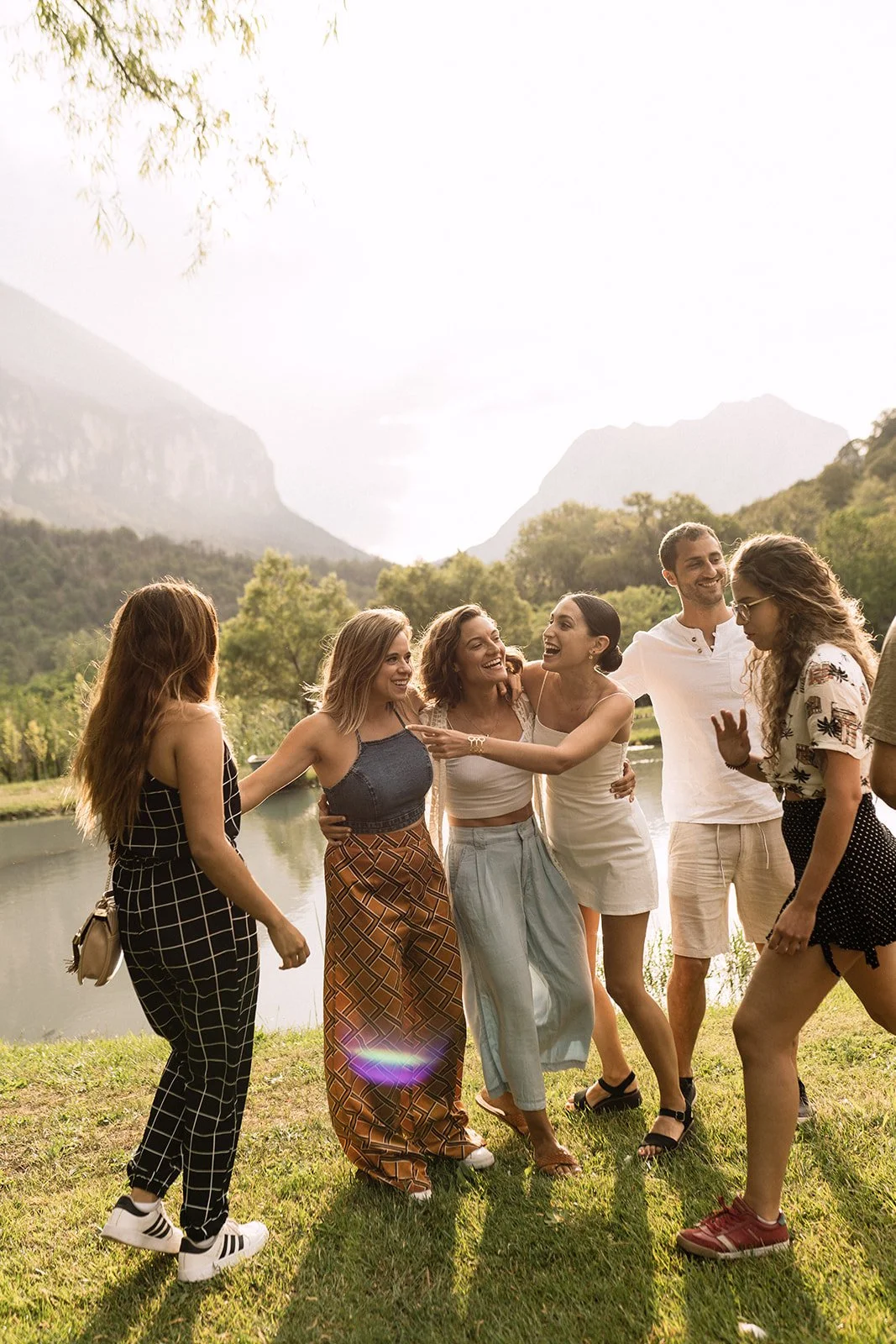 Grupo de amigos riendo y conviviendo al aire libre cerca de un río, con montañas y árboles en el fondo.