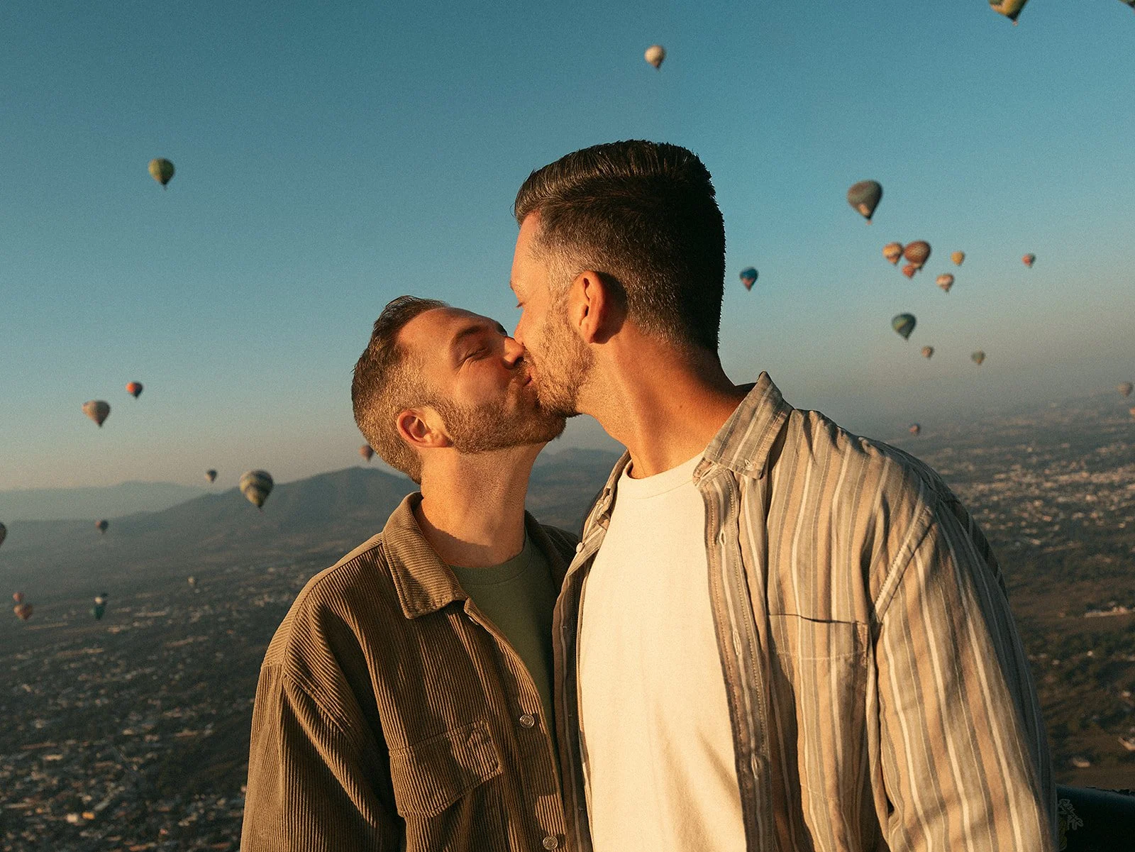 Dos hombres se besan en la mejilla frente a un cielo con globos aerostáticos y montañas al fondo durante el atardecer.