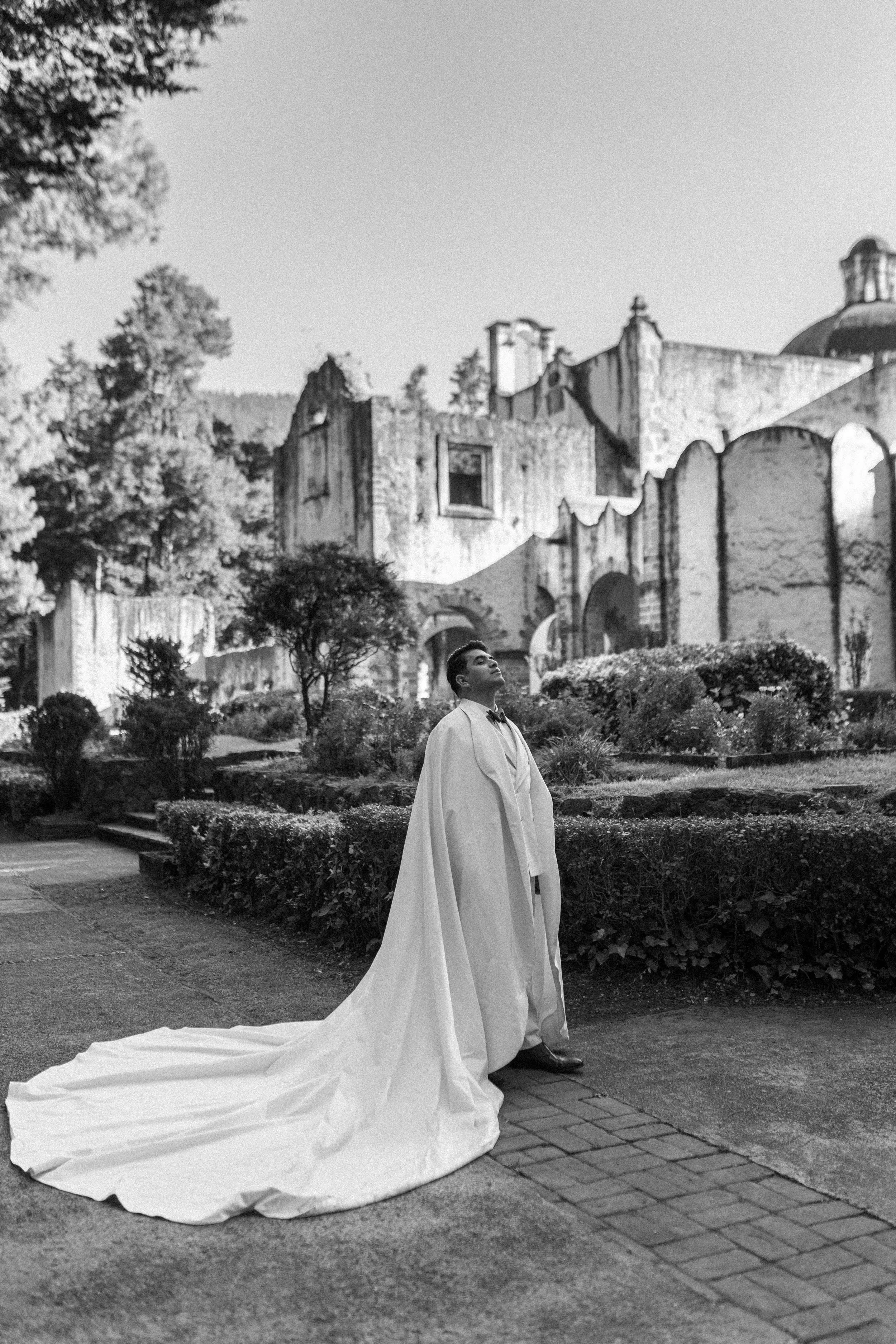 Un hombre con un traje de boda y manto largo posa en un parque frente a unas ruinas antiguas, con árboles y arbustos a su alrededor.
