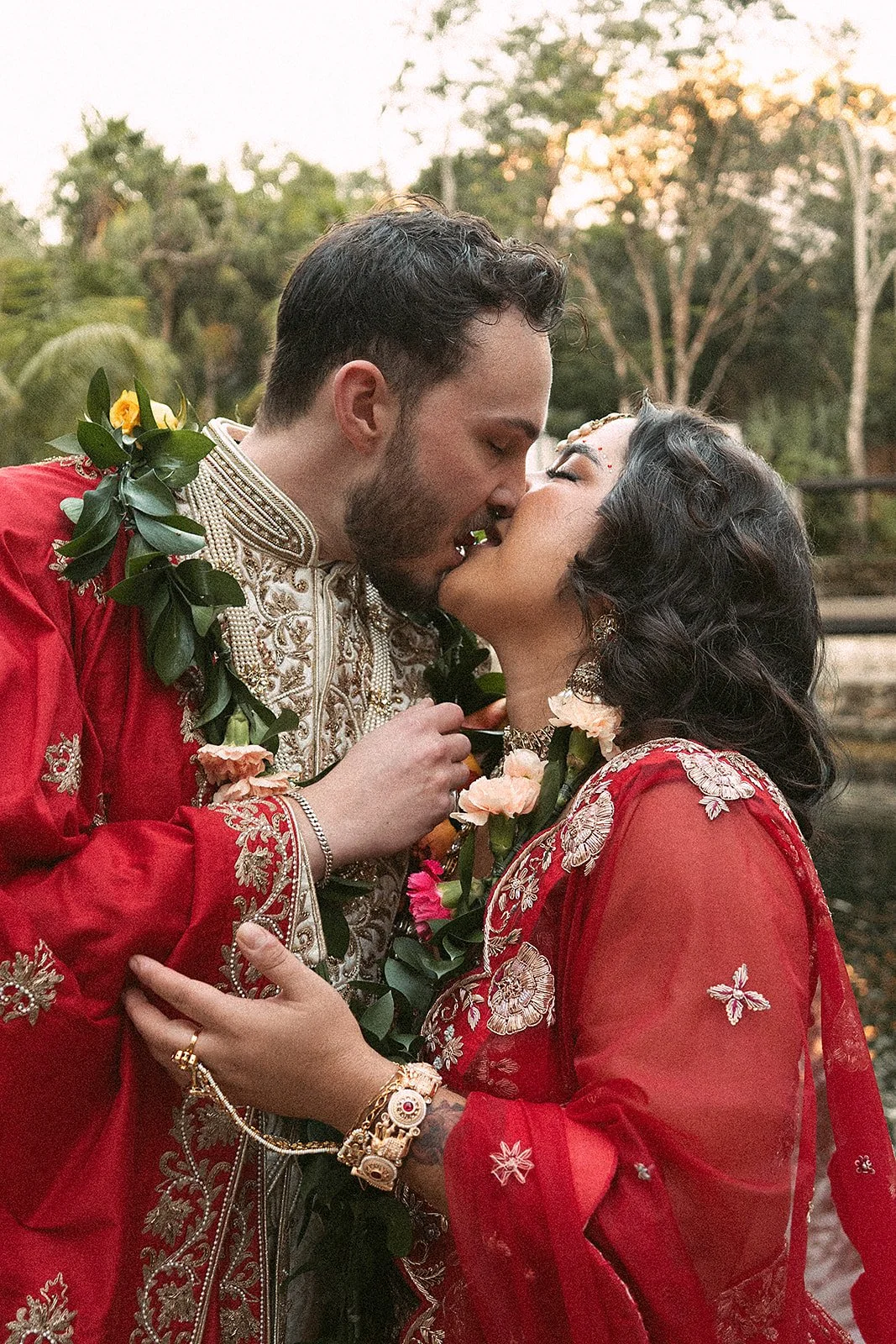 Pareja de enamorados en vestimenta tradicional, compartiendo un beso en un entorno al aire libre durante atardecer, adornados con guirnaldas de flores y joyería.