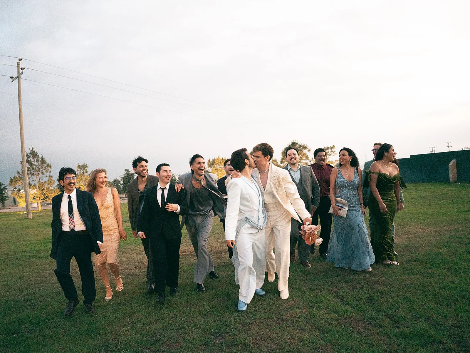 A group of people dressed in formal attire walking on grass field, two of them kissing in the center, smiling and enjoying the moment.