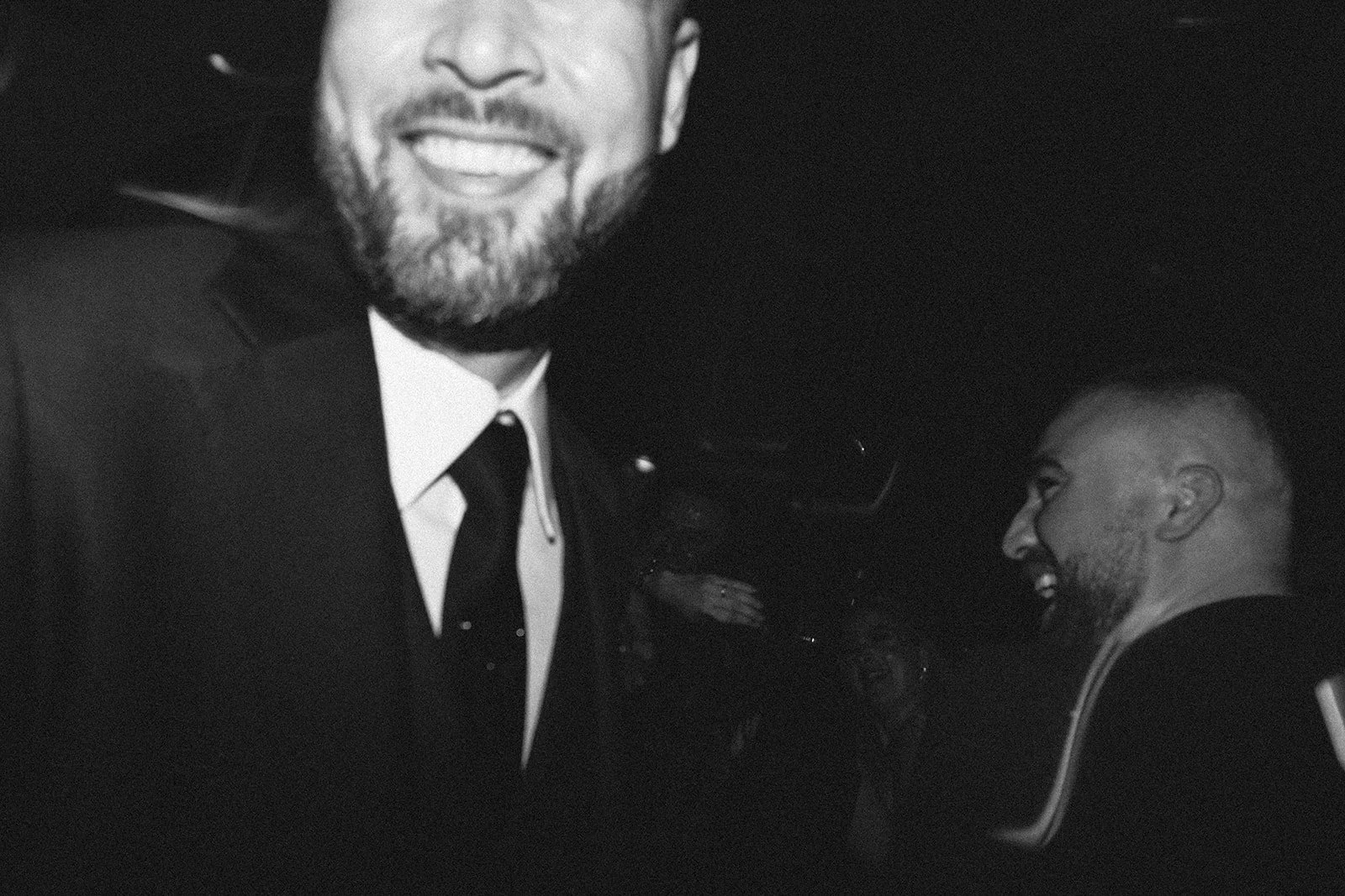 Dos hombres sonriendo en un entorno oscuro; uno en primer plano y otro en el fondo, ambos con barba y vestidos formalmente.