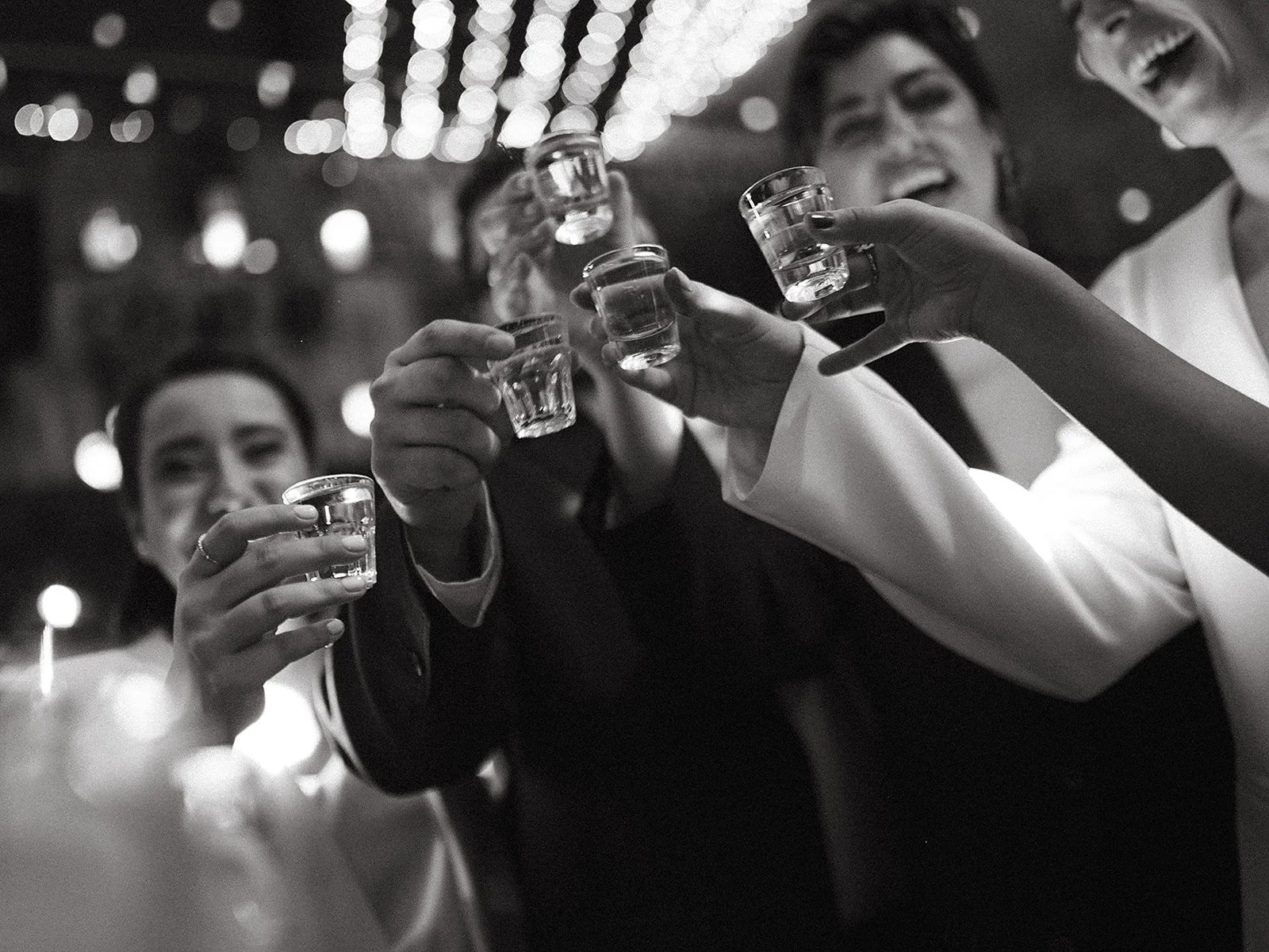 Grupo de amigos brindando con shots en una celebración, sonriendo y disfrutando en un ambiente festivo con luces en el fondo.