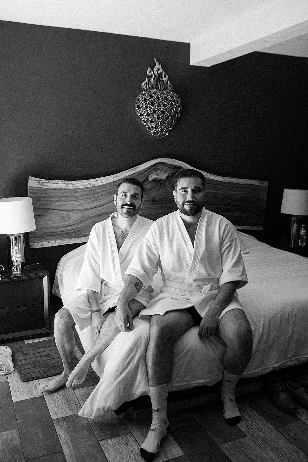 Dos hombres en bata de baño sentados en una cama en una habitación de hotel, en una imagen en escala de grises.