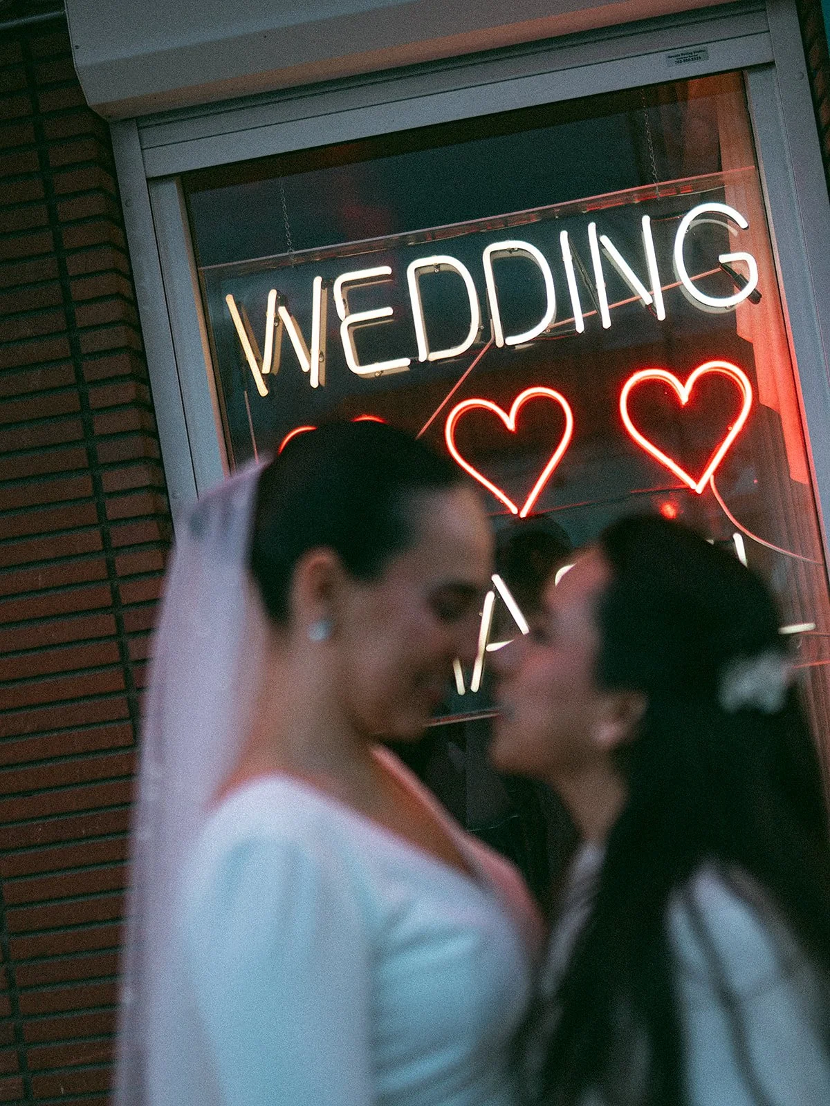 Dos novias besándose frente a un cartel de neón que dice 'WEDDING' y tiene corazones rojos de neón.