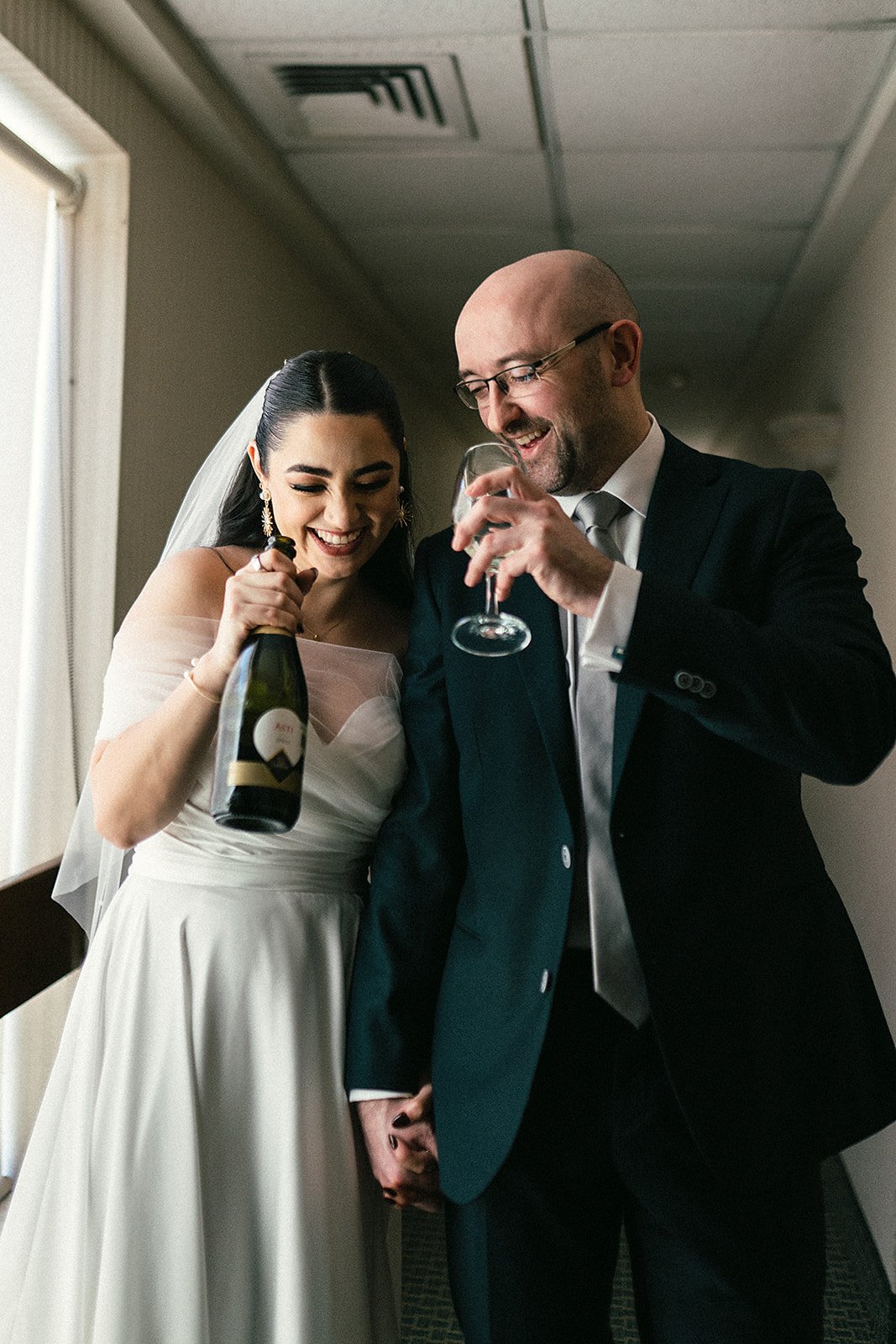 Una pareja de novios en su boda celebrando, la novia en vestido blanco con velo y el novio en traje negro, brindando con una copa de vino y la novia sosteniendo una botella de champagne.