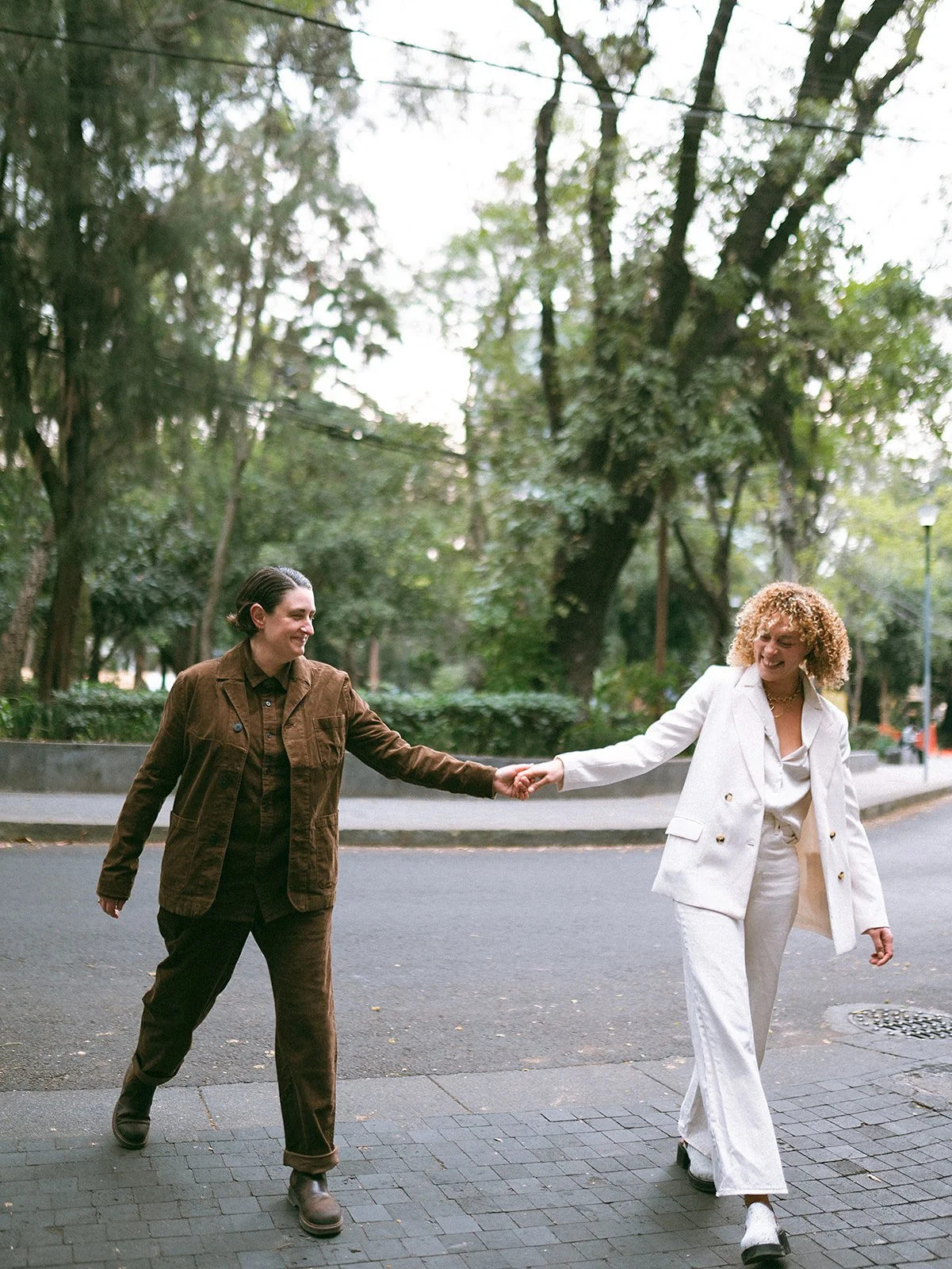 Dos mujeres caminando y tomándose de las manos en un parque urbano con árboles verdes y banca, vestidas con ropa formal, una con traje marrón y otra con traje blanco, ambas sonrientes.