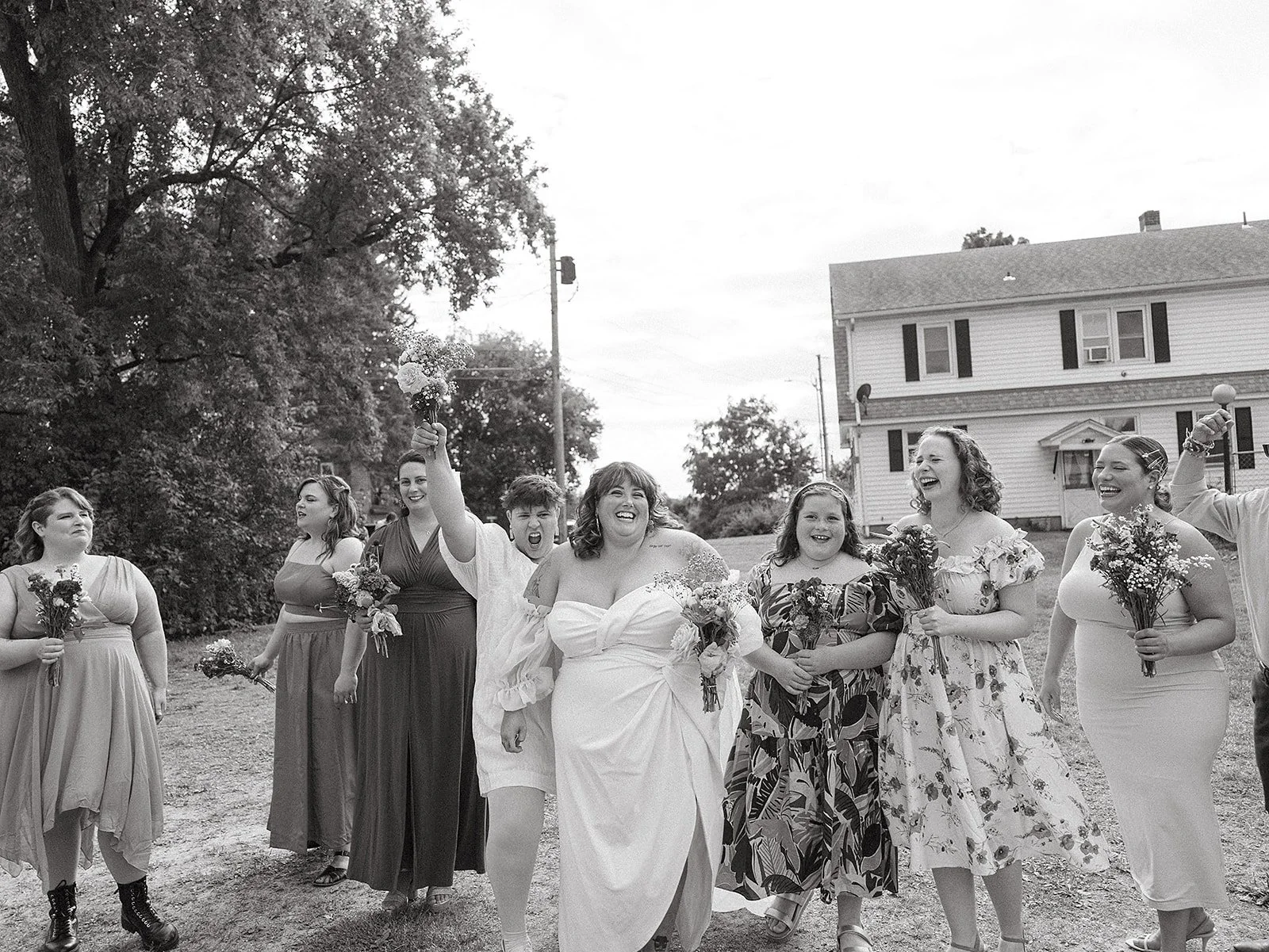 Grupo de mujeres en un evento al aire libre, algunas sosteniendo flores, todas sonriendo y vestidas con ropa casual y de fiesta, en un ambiente de patio con árbol y casa en el fondo.