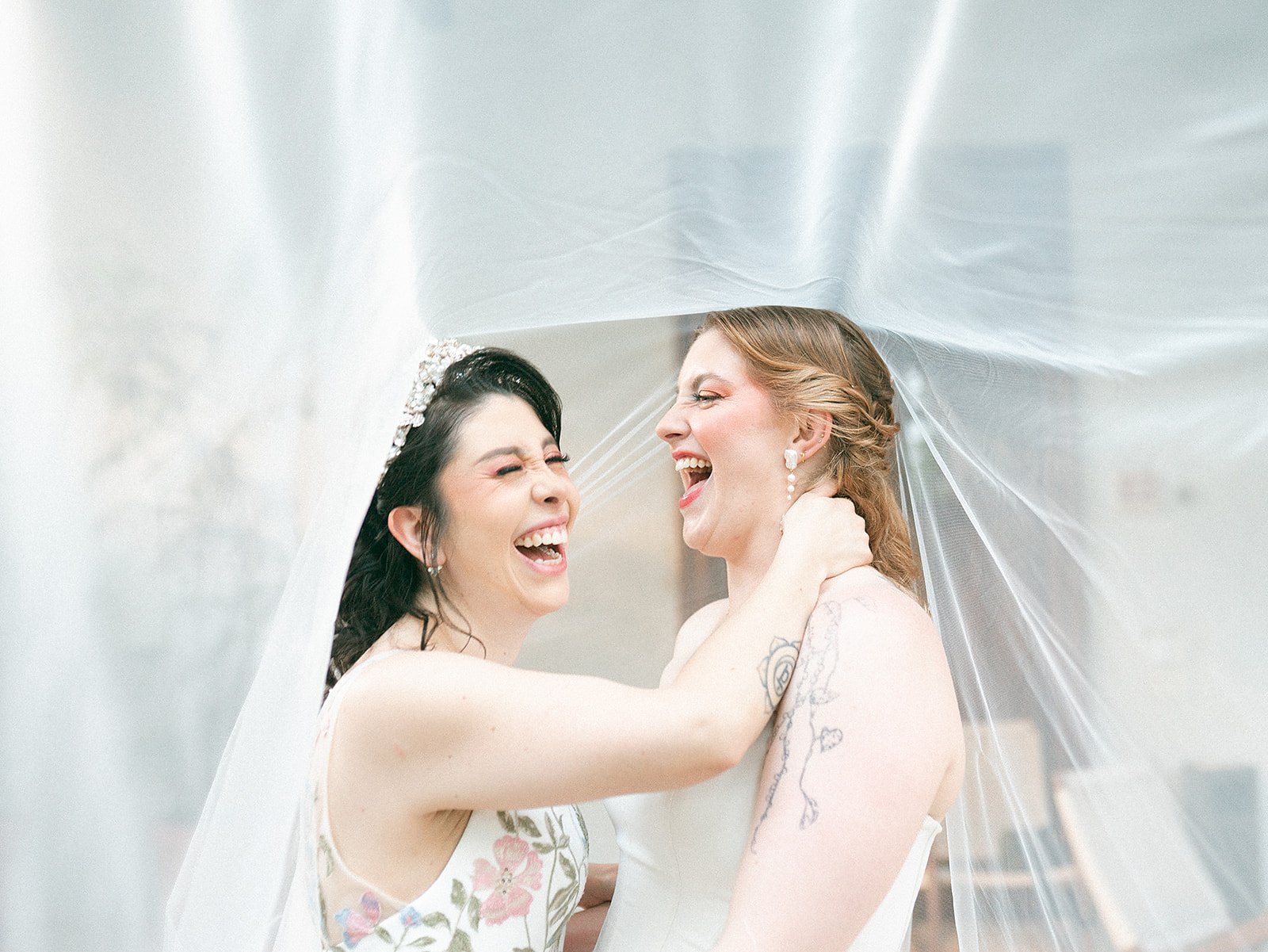 Dos mujeres en boda riendo juntas, abrazándose, con velo de novia visible.