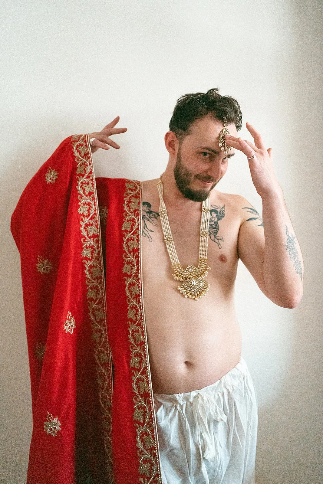Hombre con barba y tatuajes, vestido con joyas doradas y pañuelo rojo bordado, posando con una mano en la frente en un fondo simple.