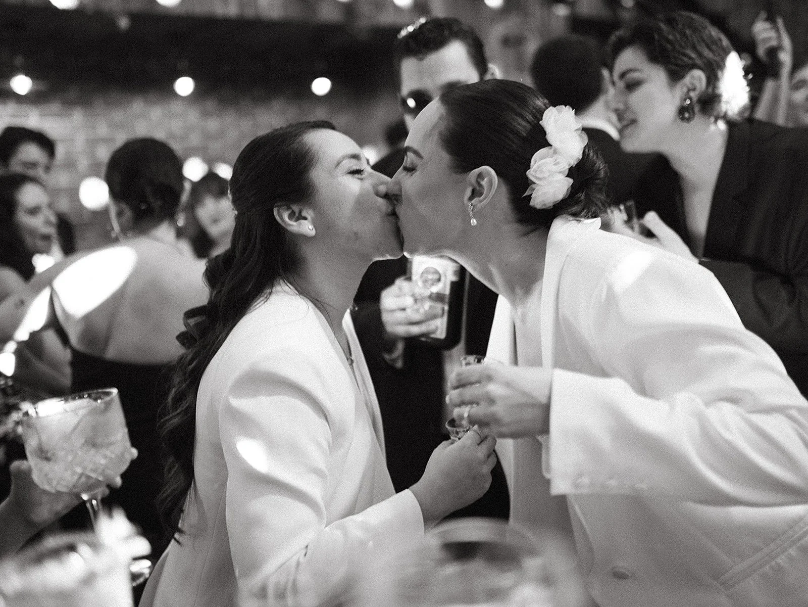 Dos mujeres se besan en un evento social, rodeadas de otras personas que celebran, en un ambiente elegante y festivo.