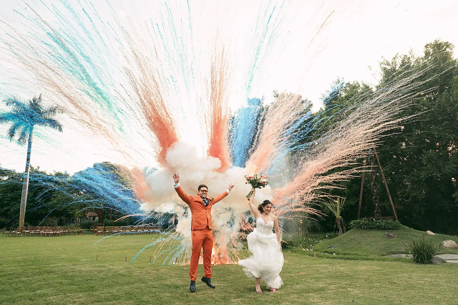 Una pareja de recién casados celebrando en un exterior con fuegos artificiales en el fondo. El hombre lleva un traje naranja y la mujer un vestido de novia, ella sostiene un ramo de flores.