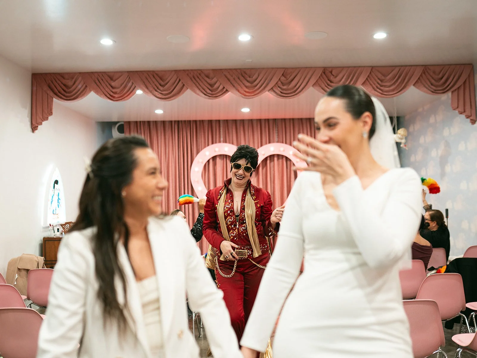 Mujeres bailando en una celebración, con decoración de corazones de luz y cortinas rosas en el fondo.