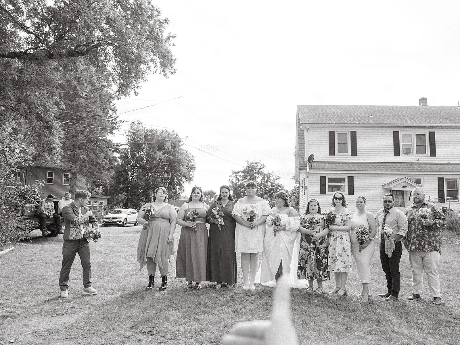Grupo de personas en una celebración al aire libre, incluyendo mujeres con vestidos y hombres con ropa formal, en un entorno con casas y árboles.