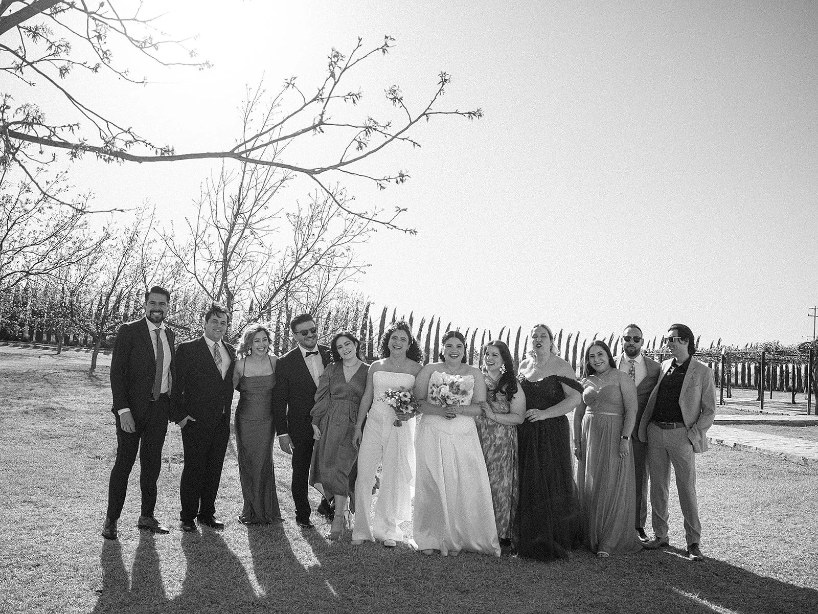 Grupo de personas vestidas con ropa formal, en un entorno al aire libre, probablemente en una boda, con árboles y cielo despejado en el fondo.