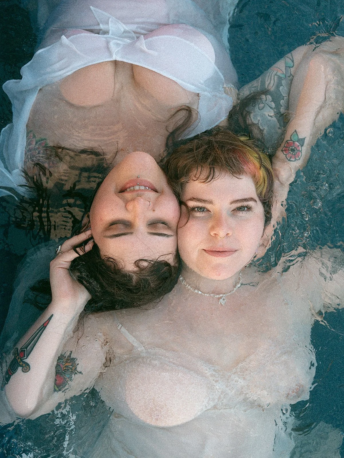 Dos jóvenes en una piscina, una con cabello rizado y la otra con cabello corto y teñido, ambas tienen tatuajes y llevan ropa de baño con accesorios. Están descansando y disfrutando del momento.