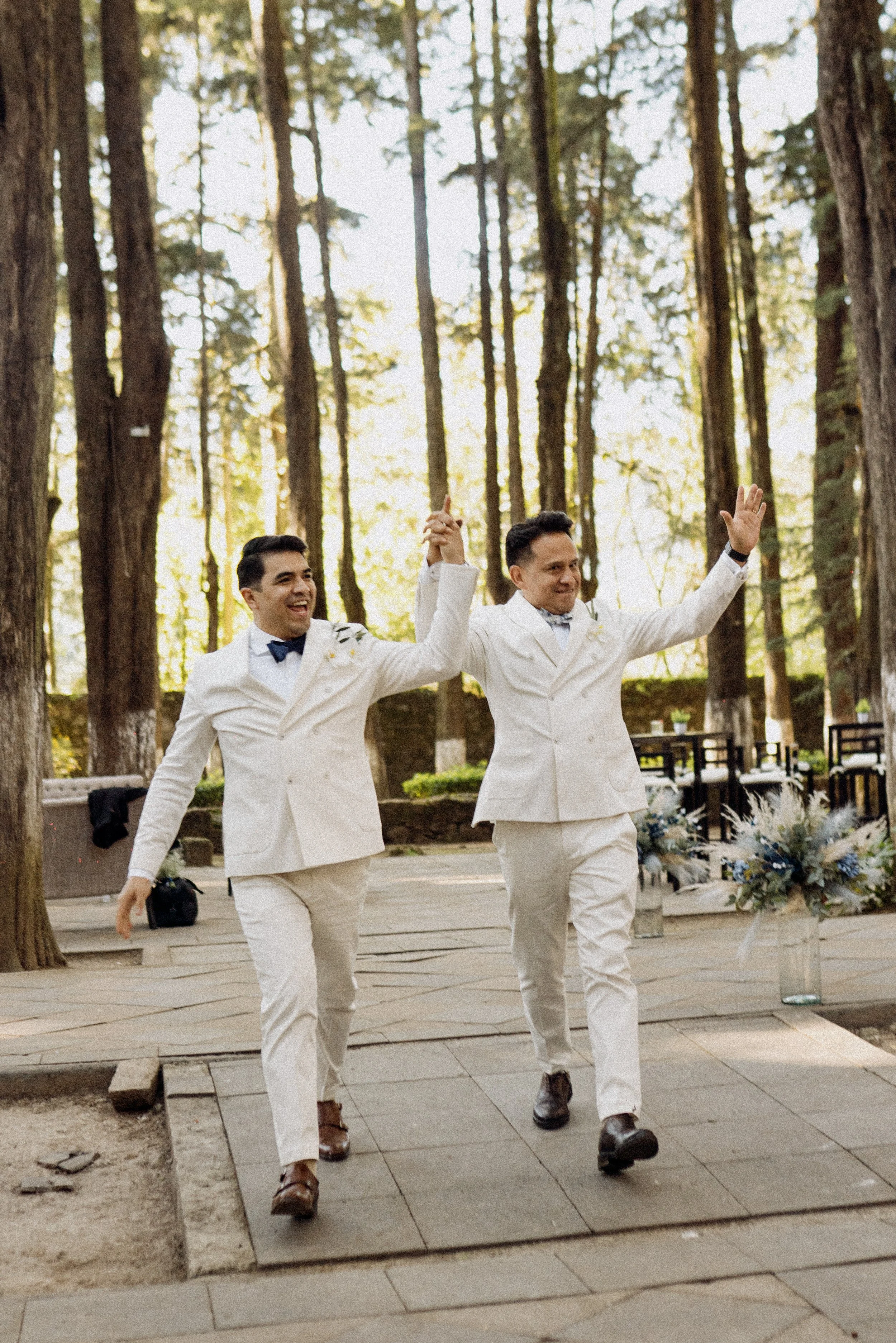 Dos hombres vestidos con trajes blancos de etiqueta elegante, uno con la cabeza levantada y el otro con la cabeza ligeramente agachada, caminando en un entorno al aire libre rodeado de árboles altos y delgados en un bosque, celebrando o disfrutando u