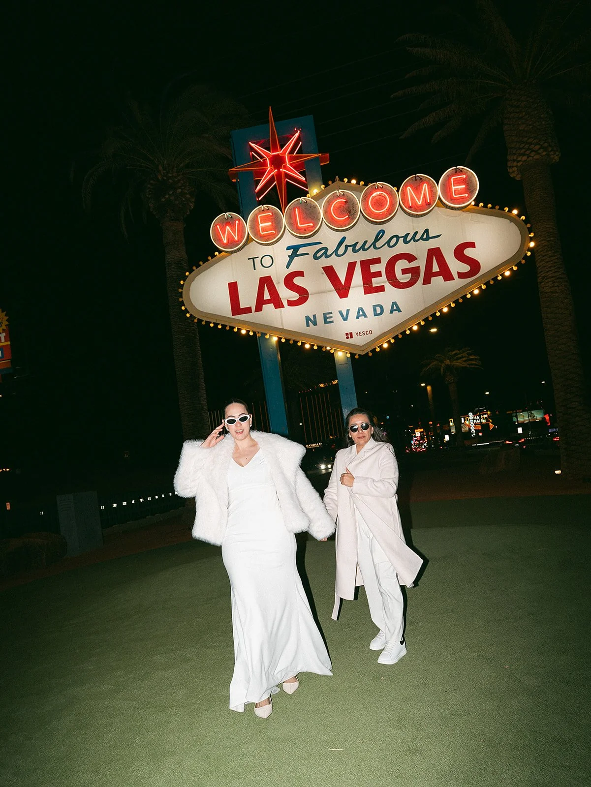 Dos mujeres con ropa elegante posando frente a la señal de bienvenida a Las Vegas, Nevada, en la noche. La señal está iluminada y decorada con un diseño brillante, acompañada de palmeras de fondo.