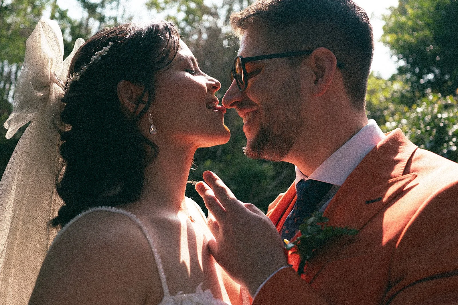 Pareja de recién casados, un hombre y una mujer, en una boda, son sonriendo y se están tocando la cara, rodeados de árboles y naturaleza.