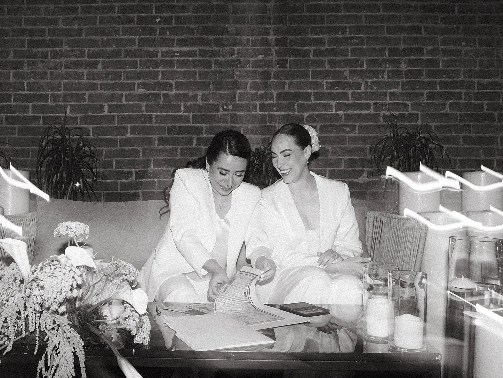 Dos mujeres con ropa formal sentadas en una mesa, sonriendo y leyendo un folleto, en un ambiente relajado con decoraciones florales y velas.