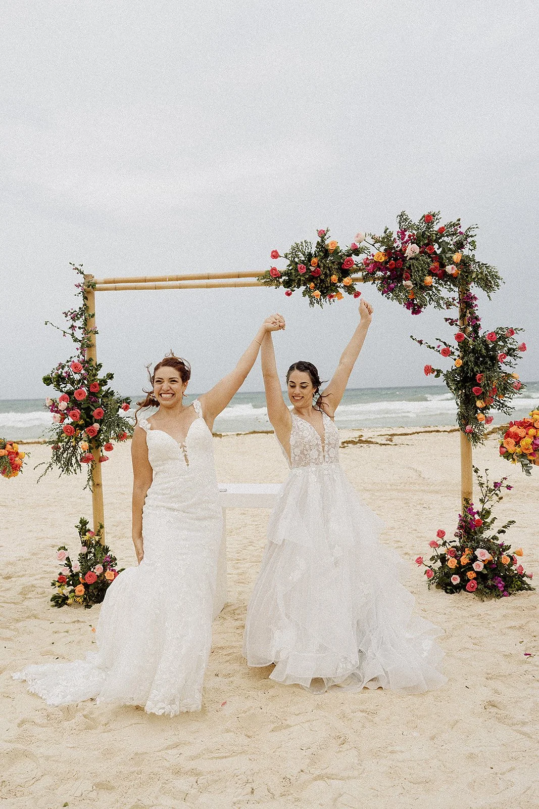 Mujeres en vestidos de novia celebrando en la playa bajo un arco de flores.