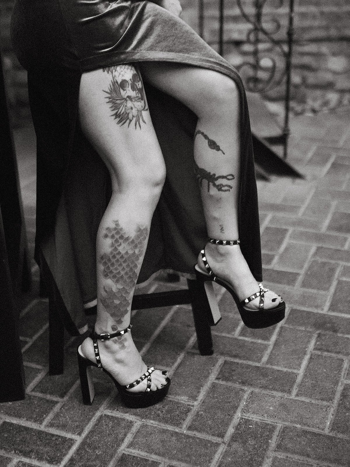 Pierna de una mujer con tatuajes, tacones altos con argollas y un vestido largo. Ella está sentada en una silla de madera sobre un piso de ladrillos.