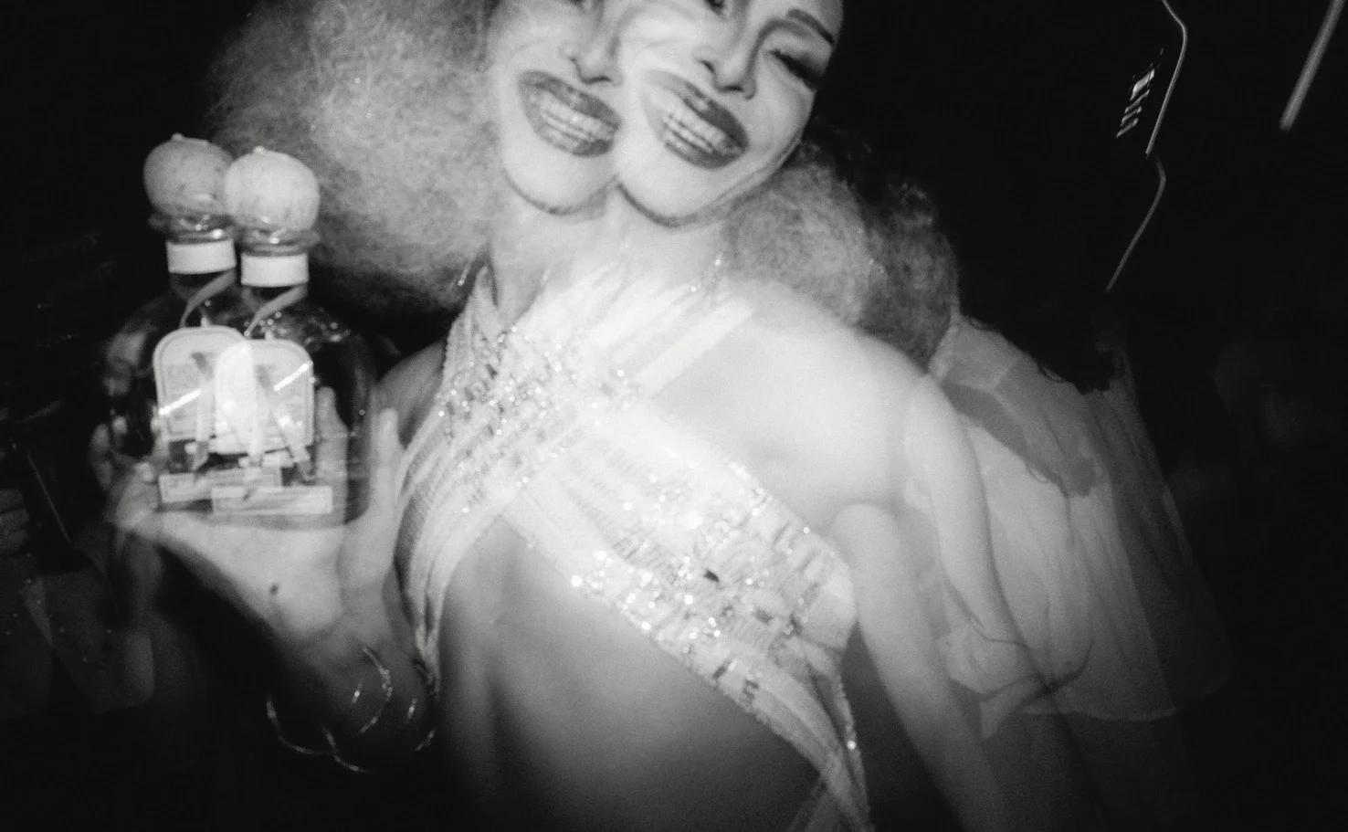 Mujer sonriendo, abrazada con varias botellas de licor, en un entorno oscuro.