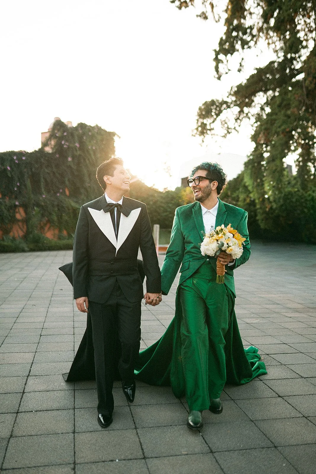 Pareja de personas en traje de boda, uno con saco verde y otro con saco negro, caminando de la mano y sonriendo en un espacio al aire libre durante el atardecer, con árboles y vegetación en el fondo.