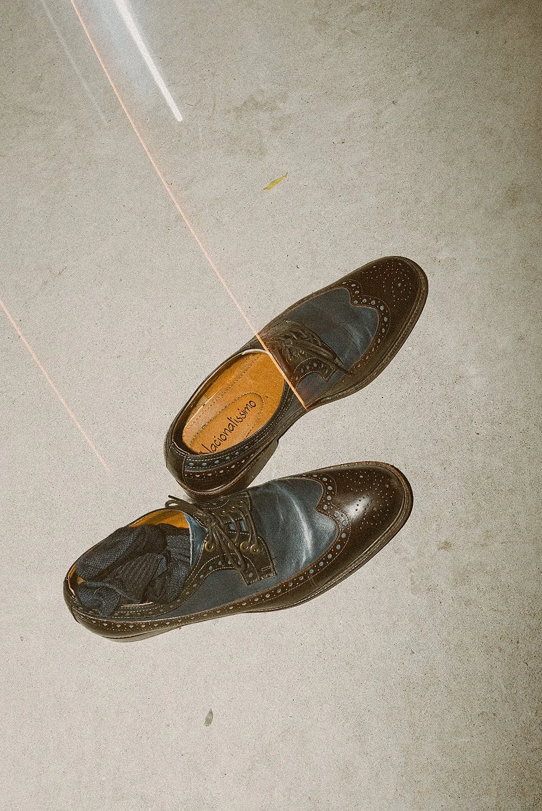 Zapatos de vestir negros y marrones con diseño clásico y tachas, con uno de ellos con calcetín y un calcetín dentro del zapato, en un suelo de cemento.