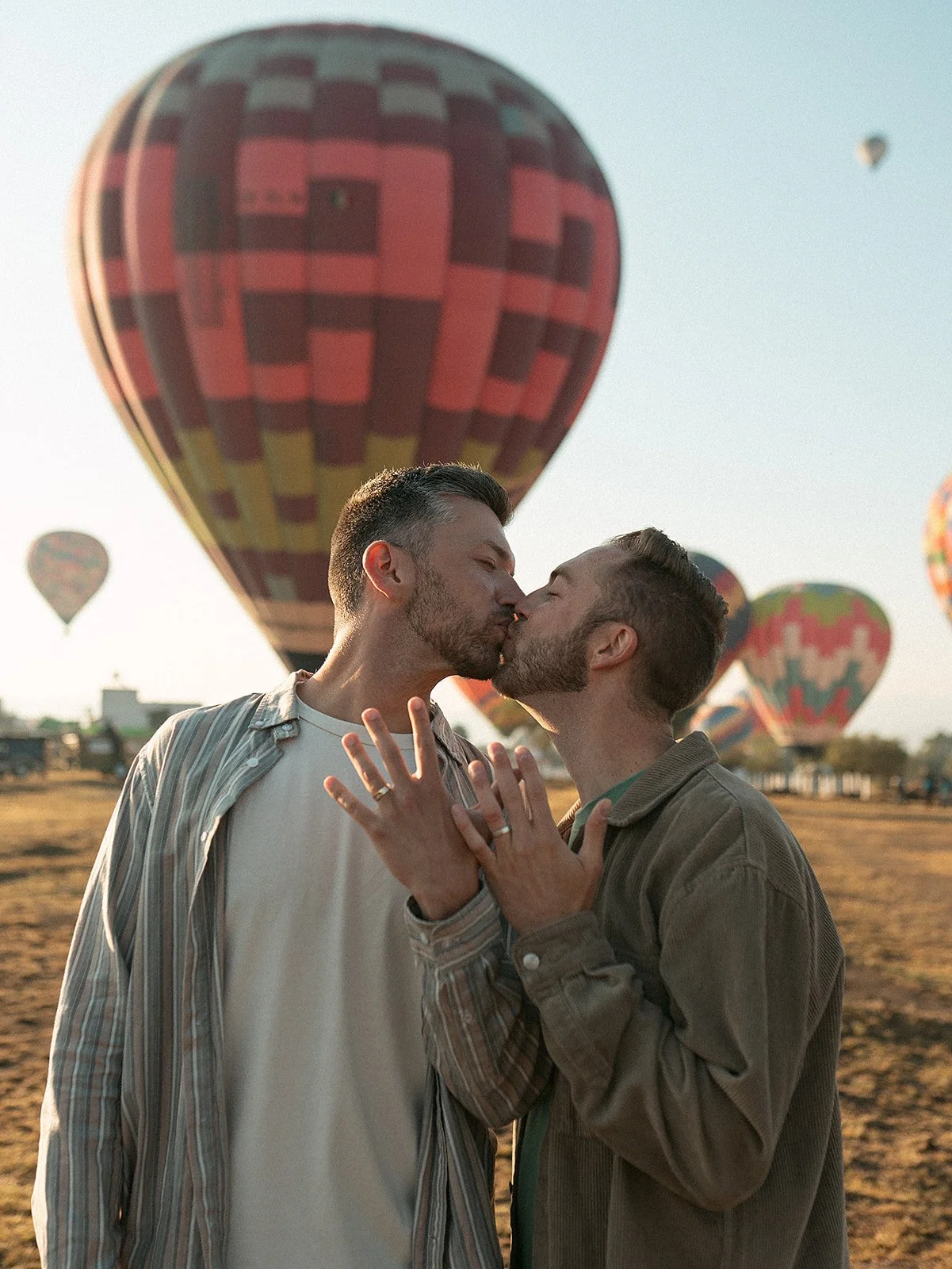 Dos hombres besándose con globos aerostáticos de fondo en un campo. 