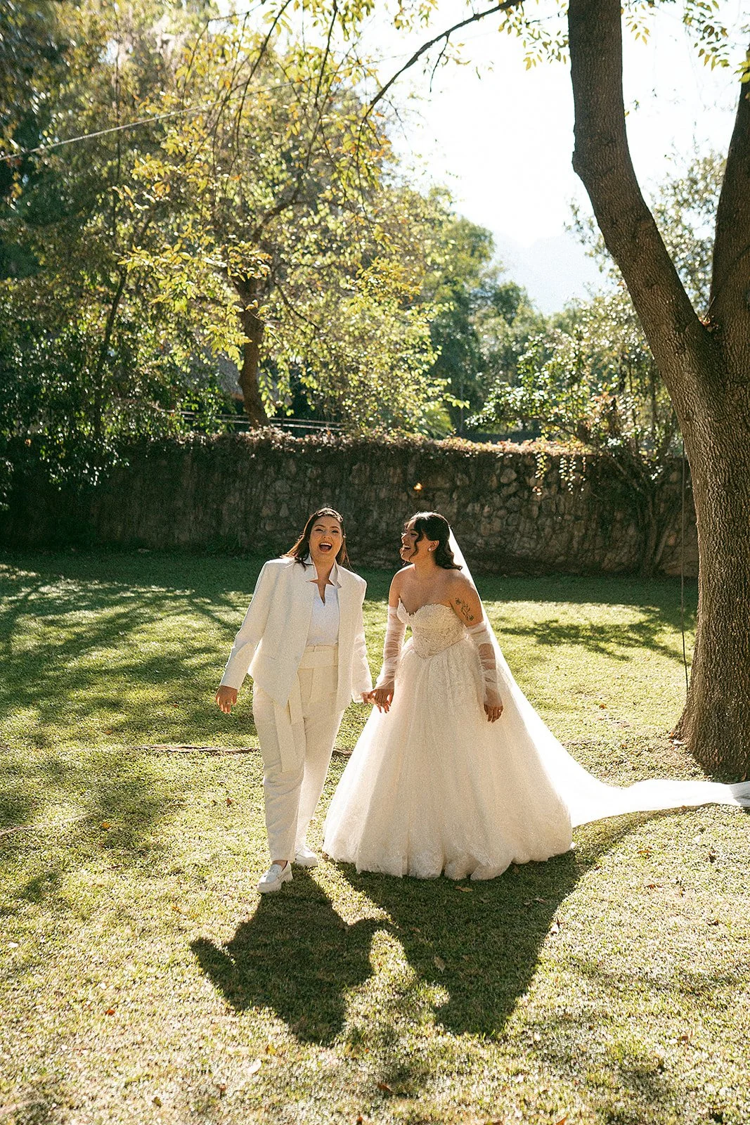 Pareja de lesbianas en vestido de novia y traje blanco, caminando y sonriendo juntas en un parque con árboles y sol.