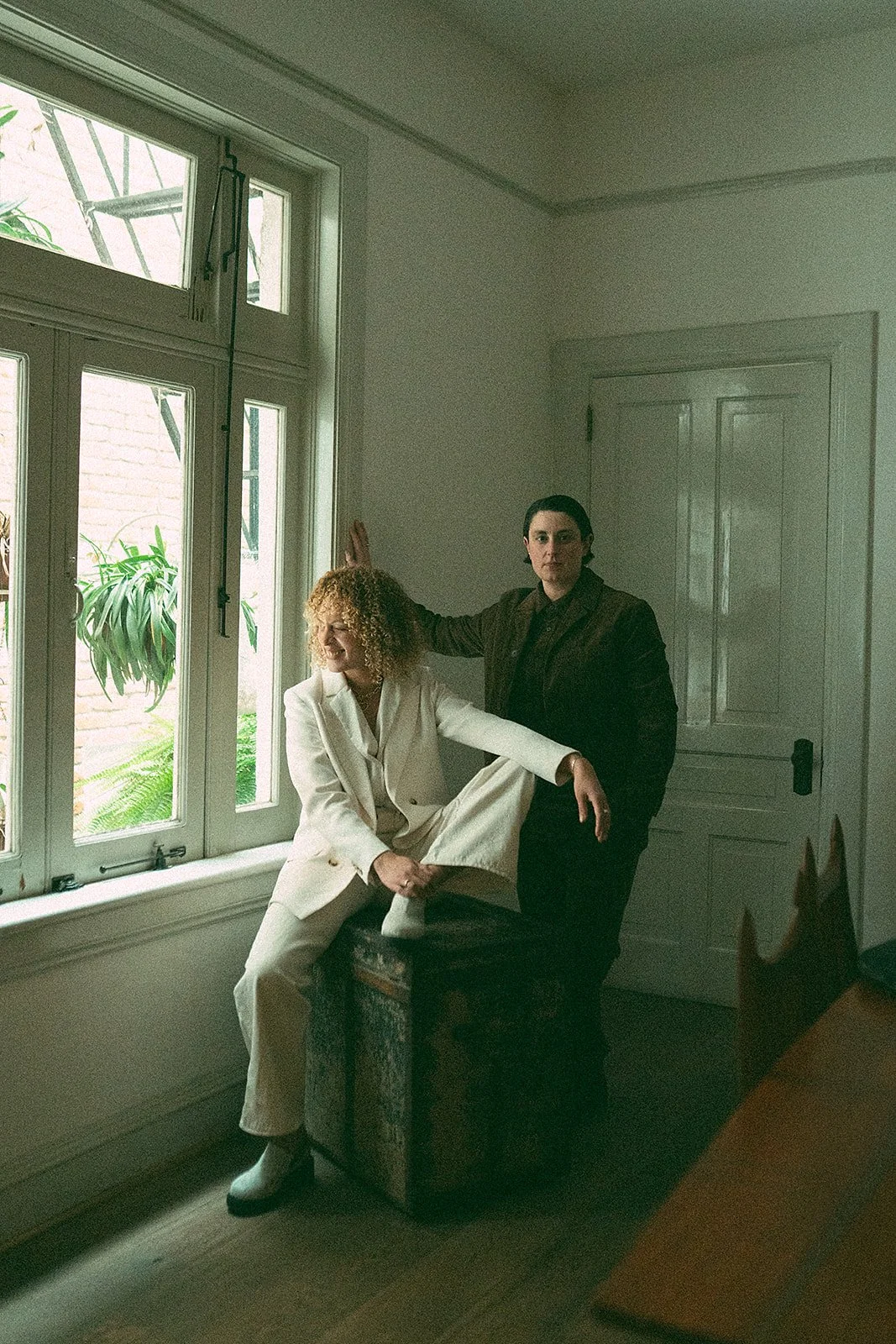 Dos personas en una habitación con ventana grande, una mujer sentada en un banco con ropa blanca y otra mujer de pie con ropa oscura, ambas posando de manera artística.