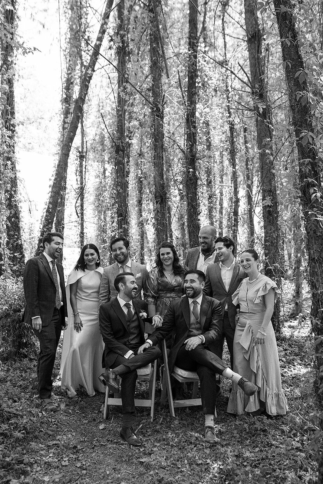 Grupo de personas en un bosque, algunas sentadas y otras de pie, en ocasión formal, todos sonriendo y disfrutando del momento.