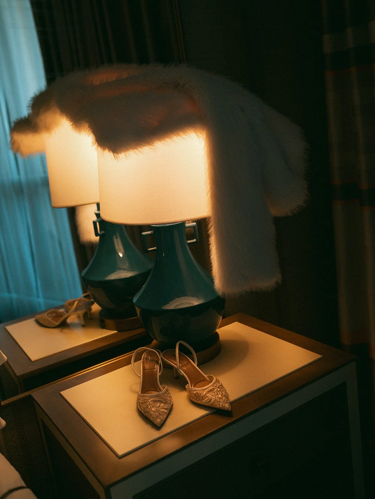 Un par de zapatos de tacón dorados con detalles de encaje y pedrería sobre una mesa cerca de dos lámparas con bases azules y pantallas blancas, en un entorno oscuro y elegante.