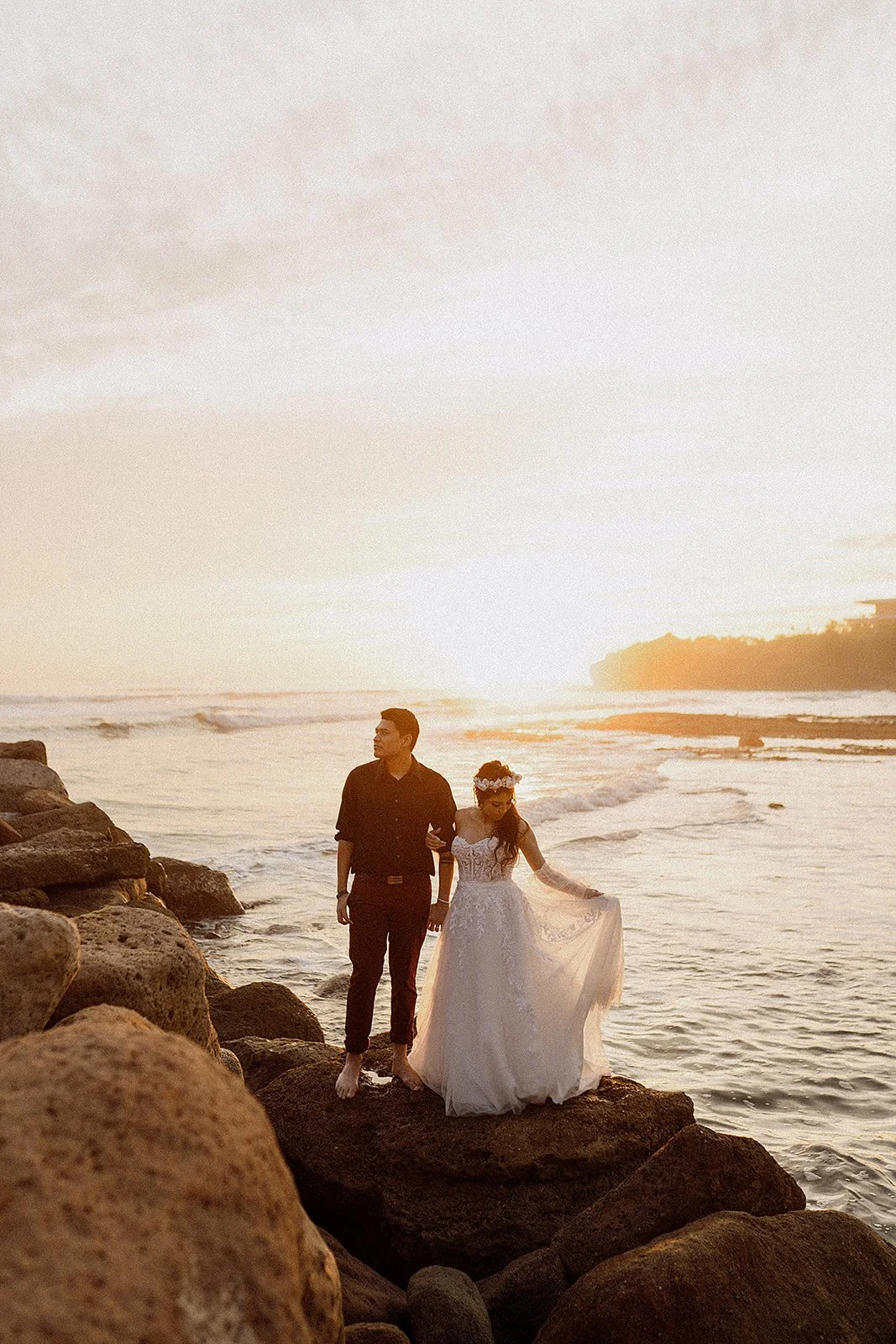 Pareja con ropa de boda caminando por las rocas en la playa al atardecer