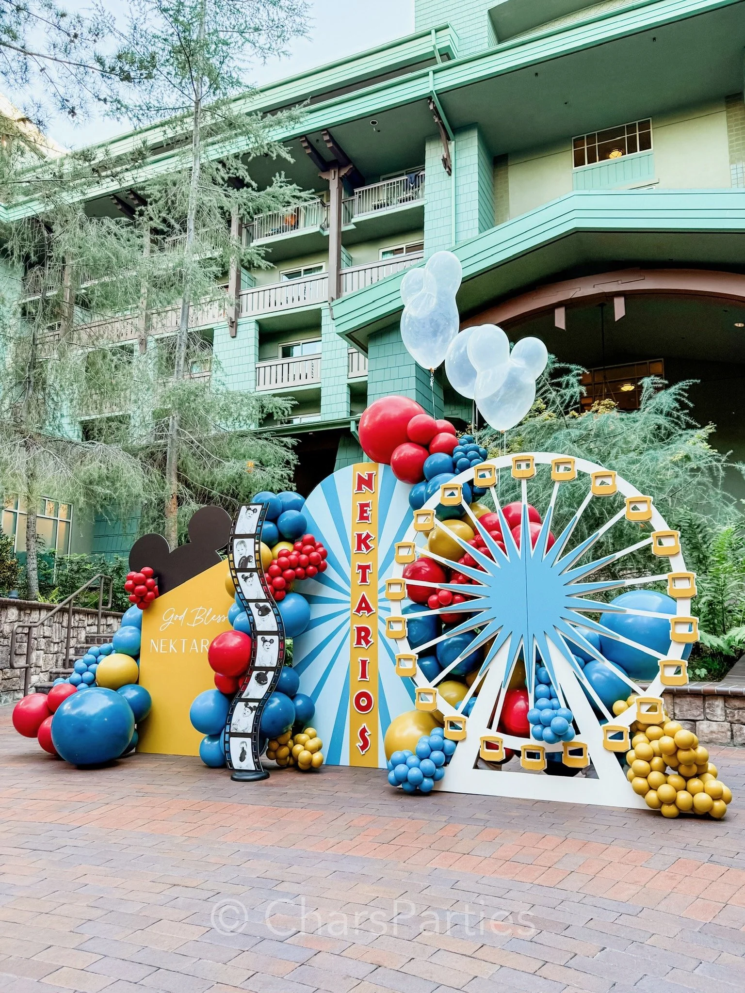 Colorful balloon display with a Ferris wheel theme, featuring red, blue, and yellow balloons, a film strip decoration, and a cutout with the text 'NEKTARIOS,' set against a green building backdrop.