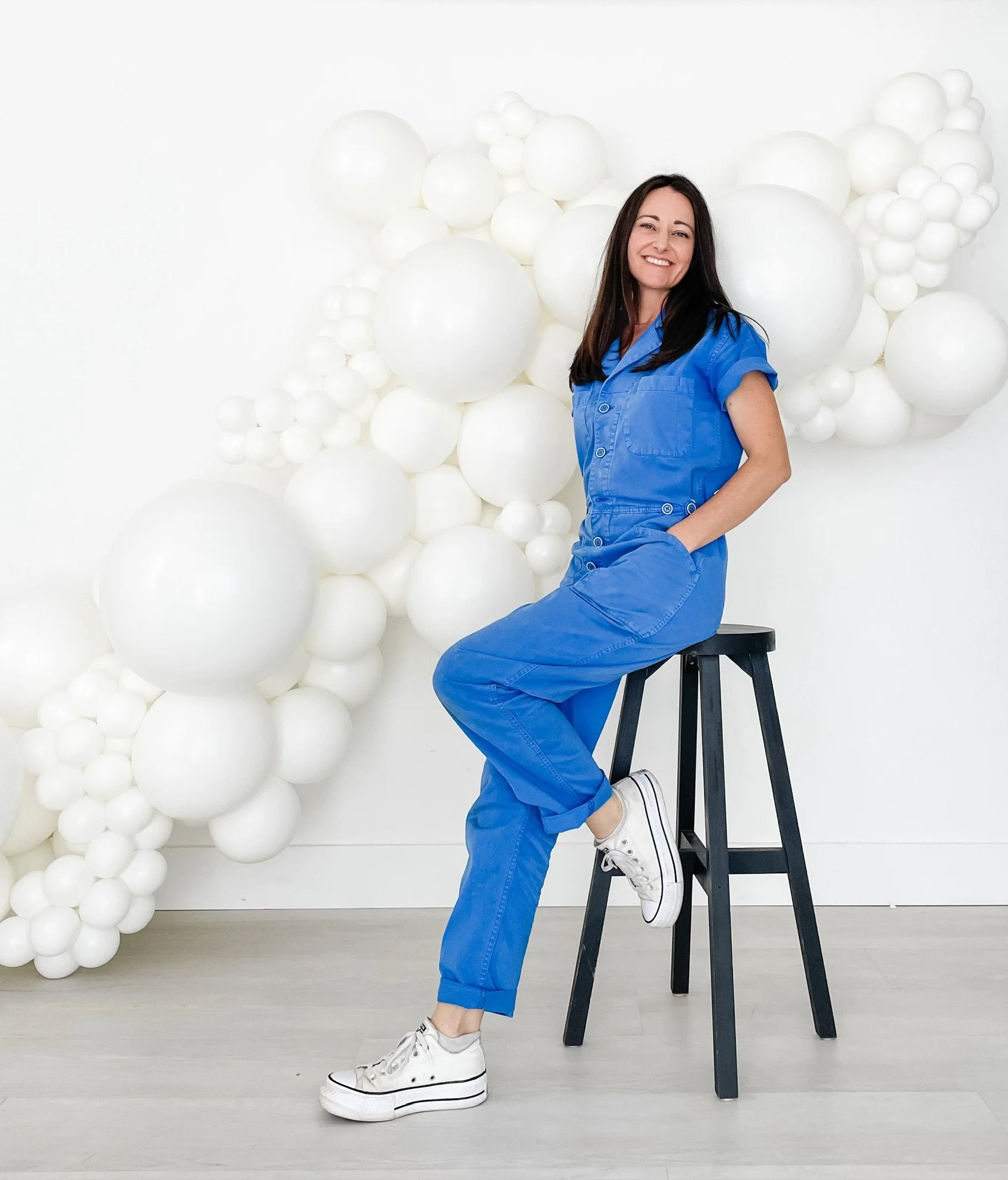 Owner/founder, Charlotte Gulley-Menchaca in blue jumpsuit sitting on stool with white balloon installation background.