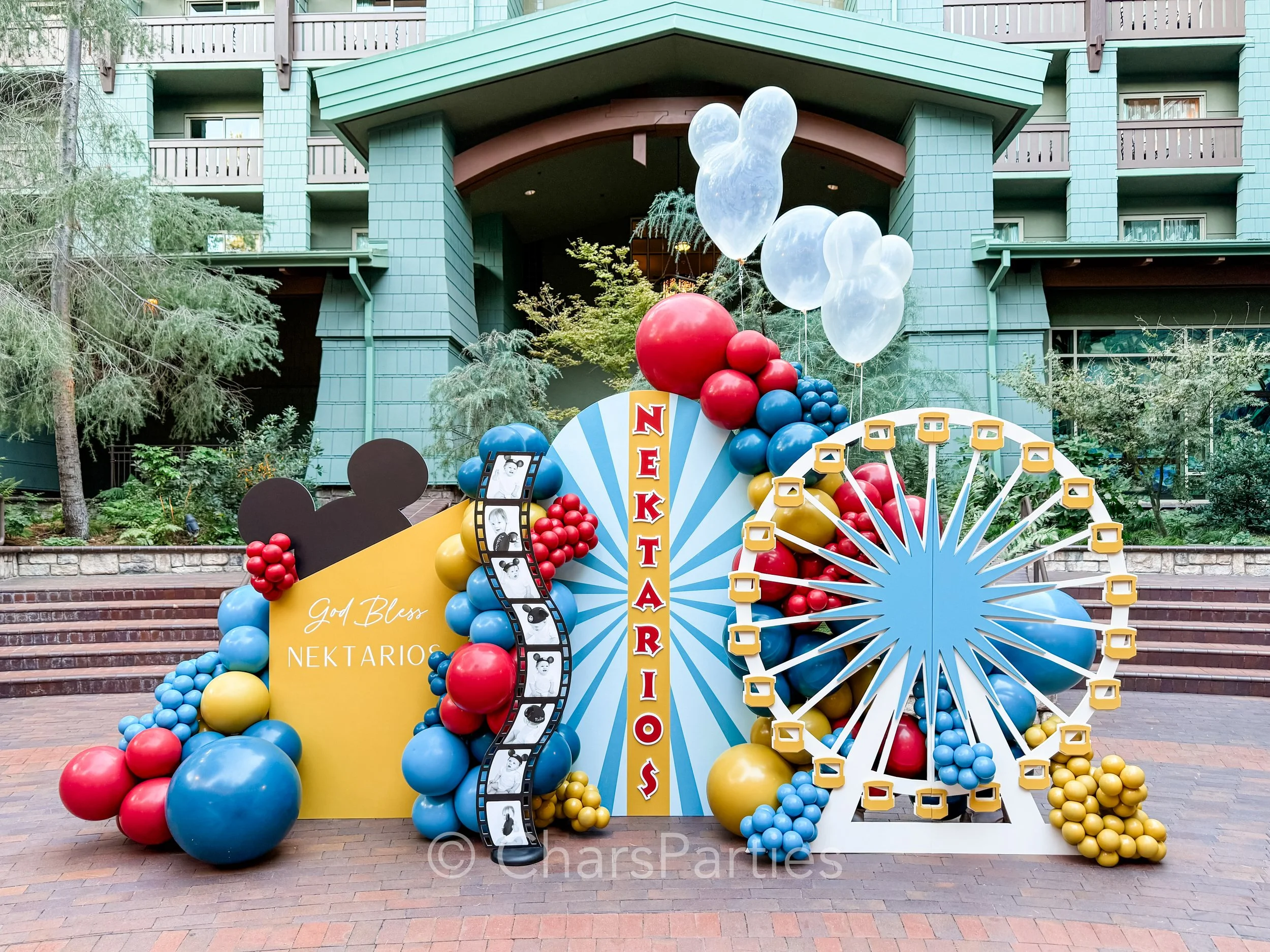 Balloon decoration display with Disney theme, featuring a Ferris wheel, Mickey Mouse silhouette, and colorful balloons in blue, red, and yellow. "God Bless Nektarios" text on a yellow backdrop, located outdoors near a green building.