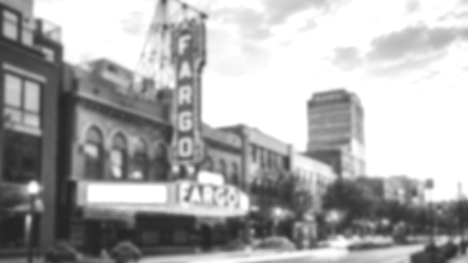Blurry black and white photo of a city street scene with a large, vertical sign reading 'FARGO' on a building, and other buildings in the background.