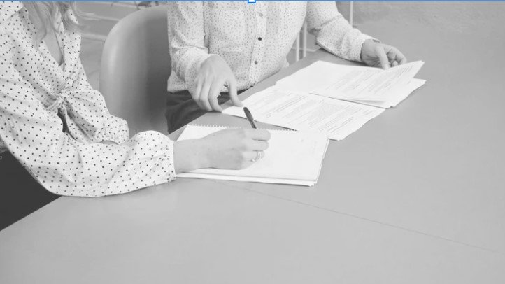 Two people sitting at a table with open papers and documents, one person pointing at a document with a pen.