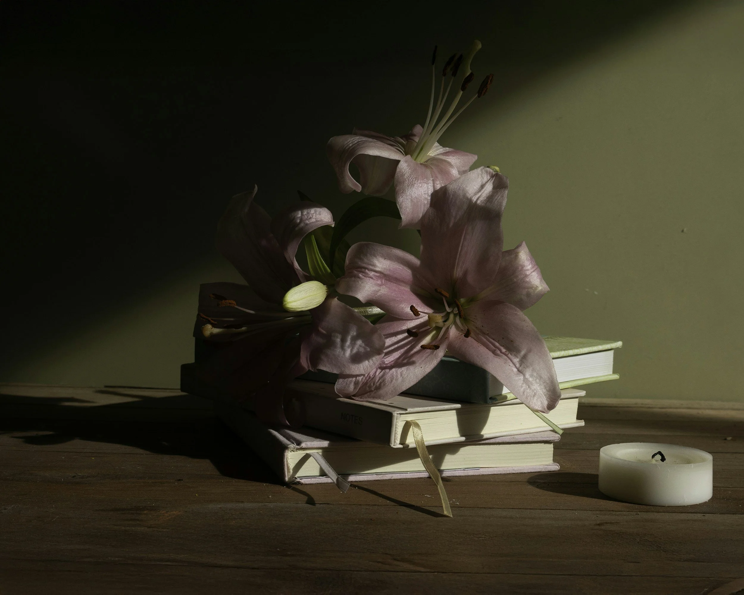 A pink lily flower resting on a stack of white notebooks on a wooden surface, with a white candle nearby and shadows cast across the scene.