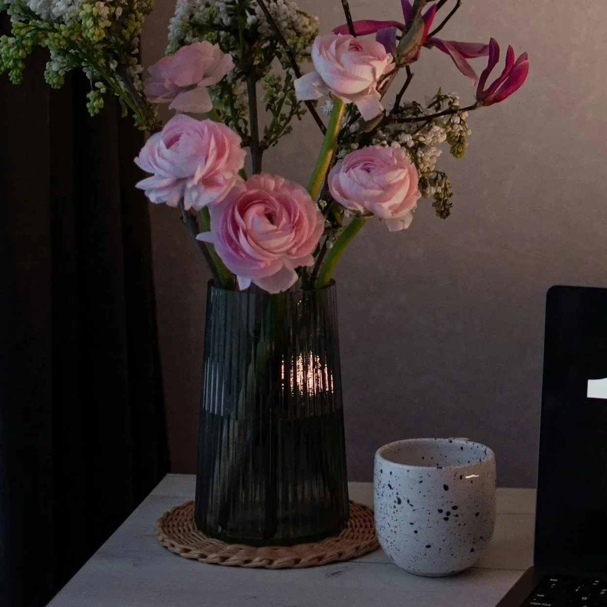 A vase with pink and white flowers on an author's desk, with a speckled white cup and part of a laptop visible.
