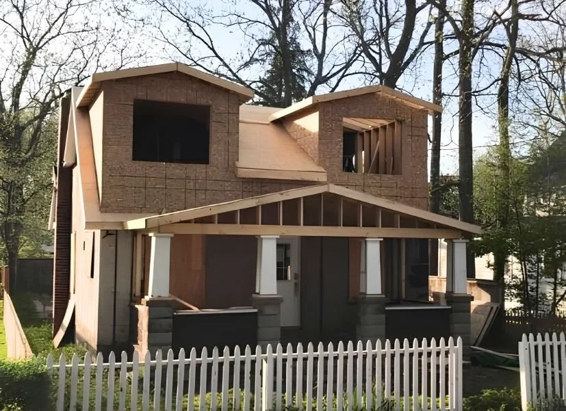 A two-story house under construction with exposed framing and brick exterior, surrounded by trees, with a white picket fence in the foreground.
