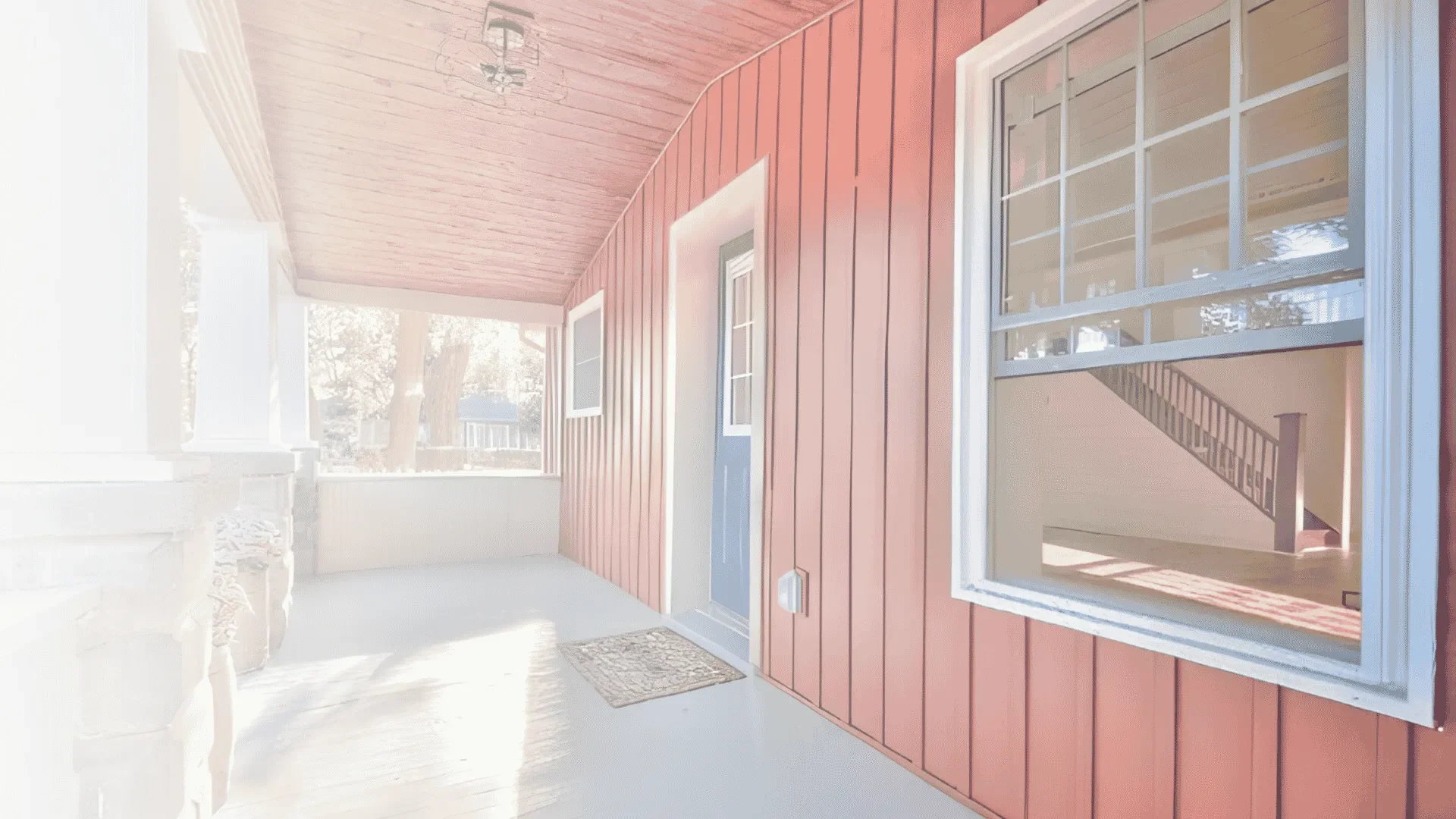 A porch with a red wooden wall, two windows, a door, and a ceiling light fixture, with a view of trees and neighboring houses outside.