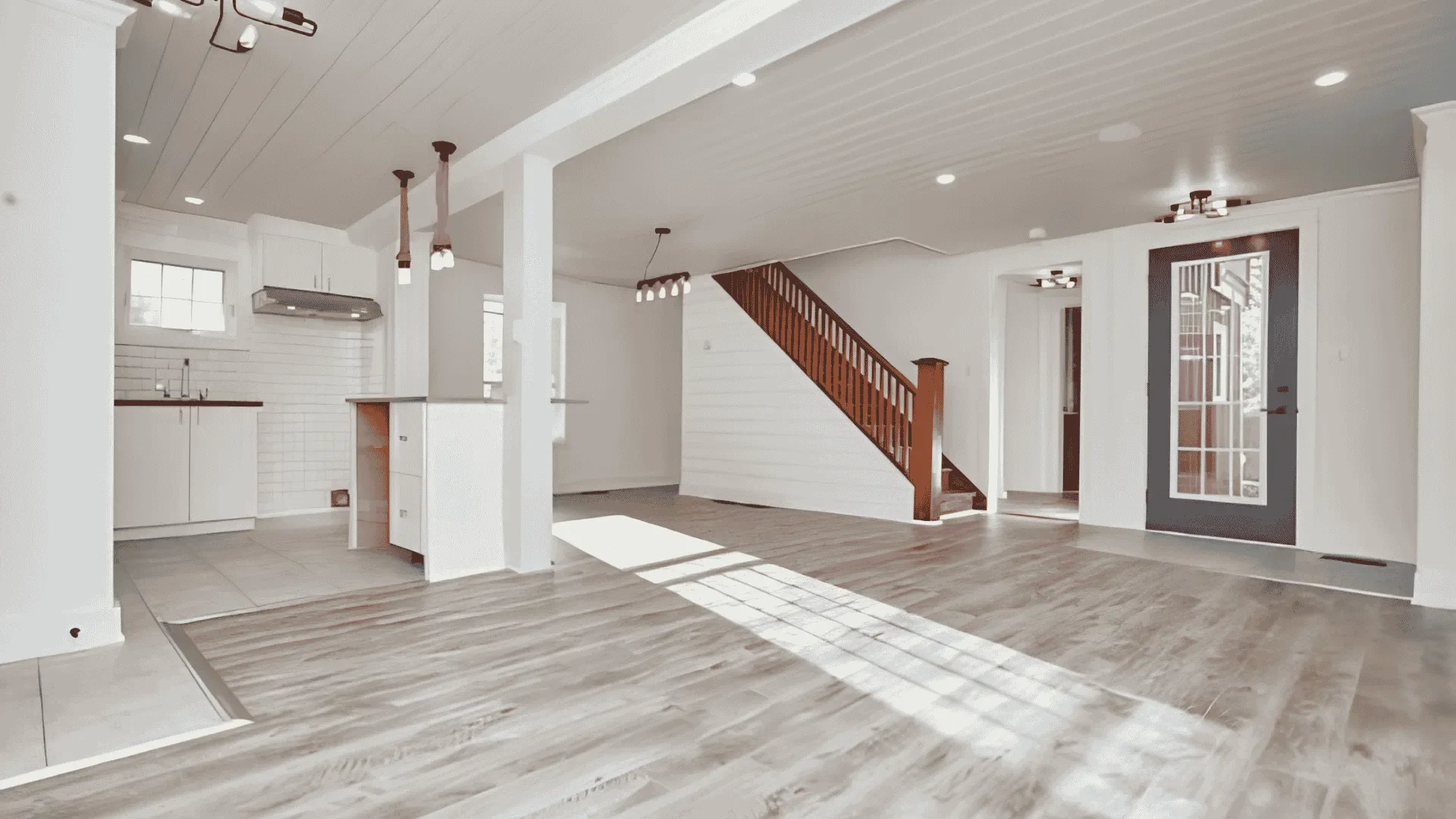 Open-concept living space with a small kitchen area, hardwood floors, white walls, staircase with wooden railing, ceiling fans, and a door leading outside.