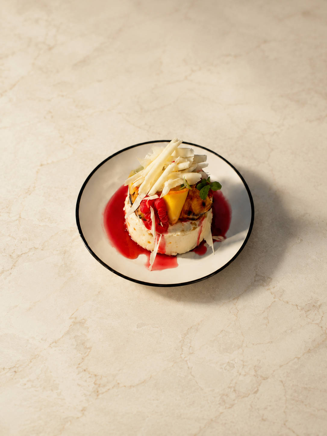 A plated dessert with white chocolate shavings, fresh fruit, and red sauce on a white plate with a black rim, placed on a marble surface.